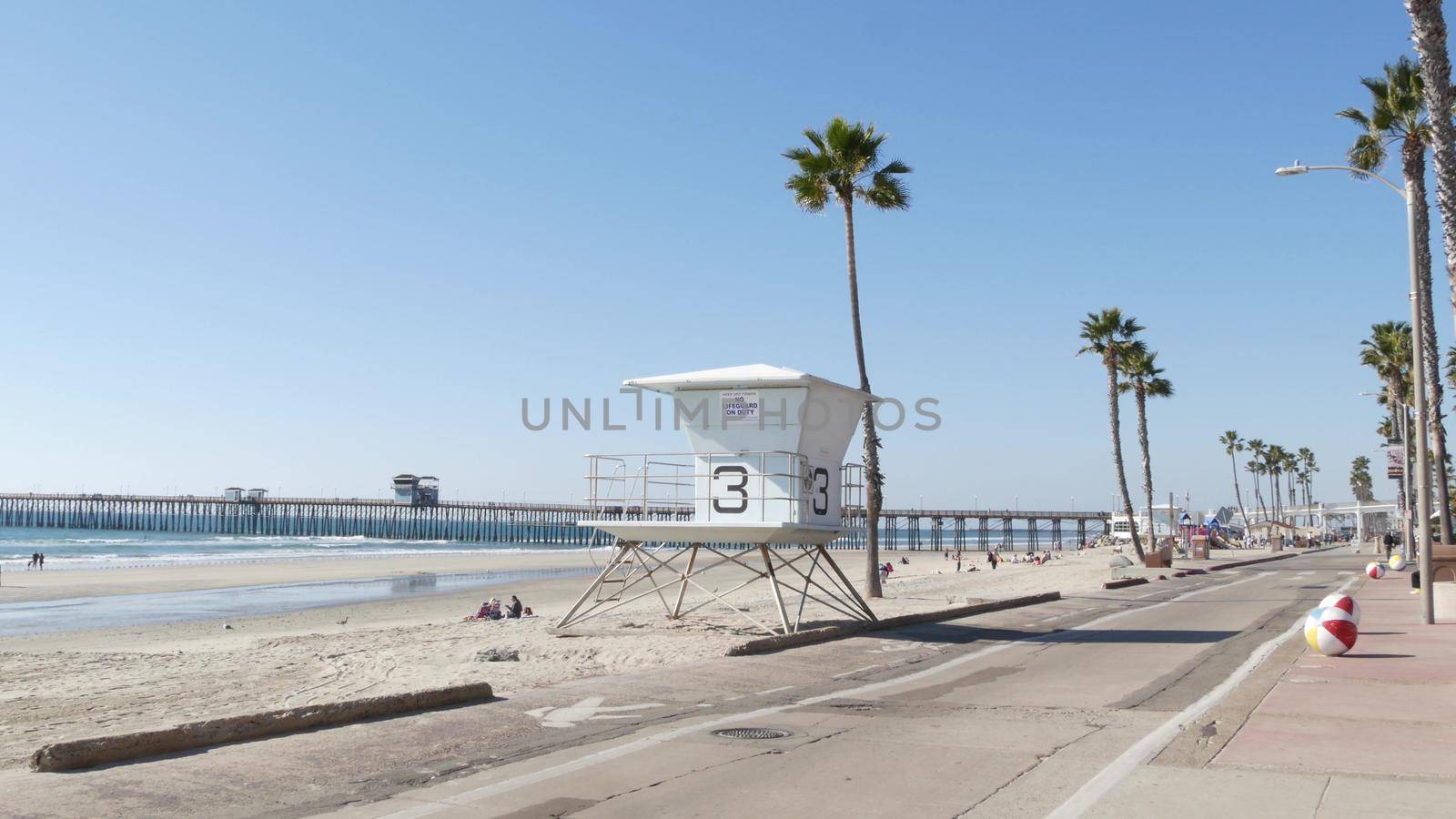 Oceanside, California USA - 8 Feb 2020: People walking, waterfront promenade near pier, pacific ocean tropical beach tourist resort with palm trees. Lifeguard tower. Disabled senior man in wheelchair.