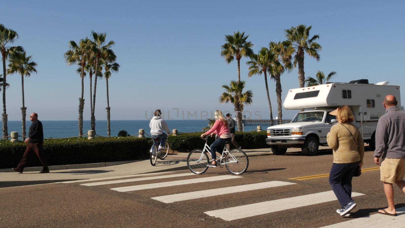 Oceanside, California USA - 8 Feb 2020: Motorhome, people riding bikes on ped crossing zebra, waterfront road. Pacific ocean tropical beach tourist resort. Palm trees on beachfront street sunny day.