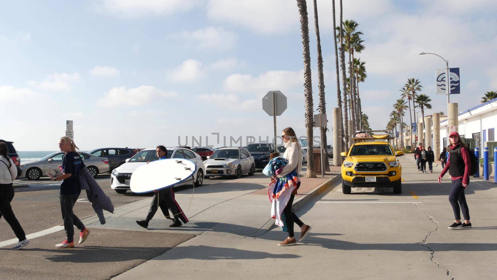 Oceanside, California USA - 8 Feb 2020: Women walking on waterfront promenade, people on beachfront boardwalk. Vacations ocean beach resort near Los Angeles. Surfers with surfboards, lifeguard car.