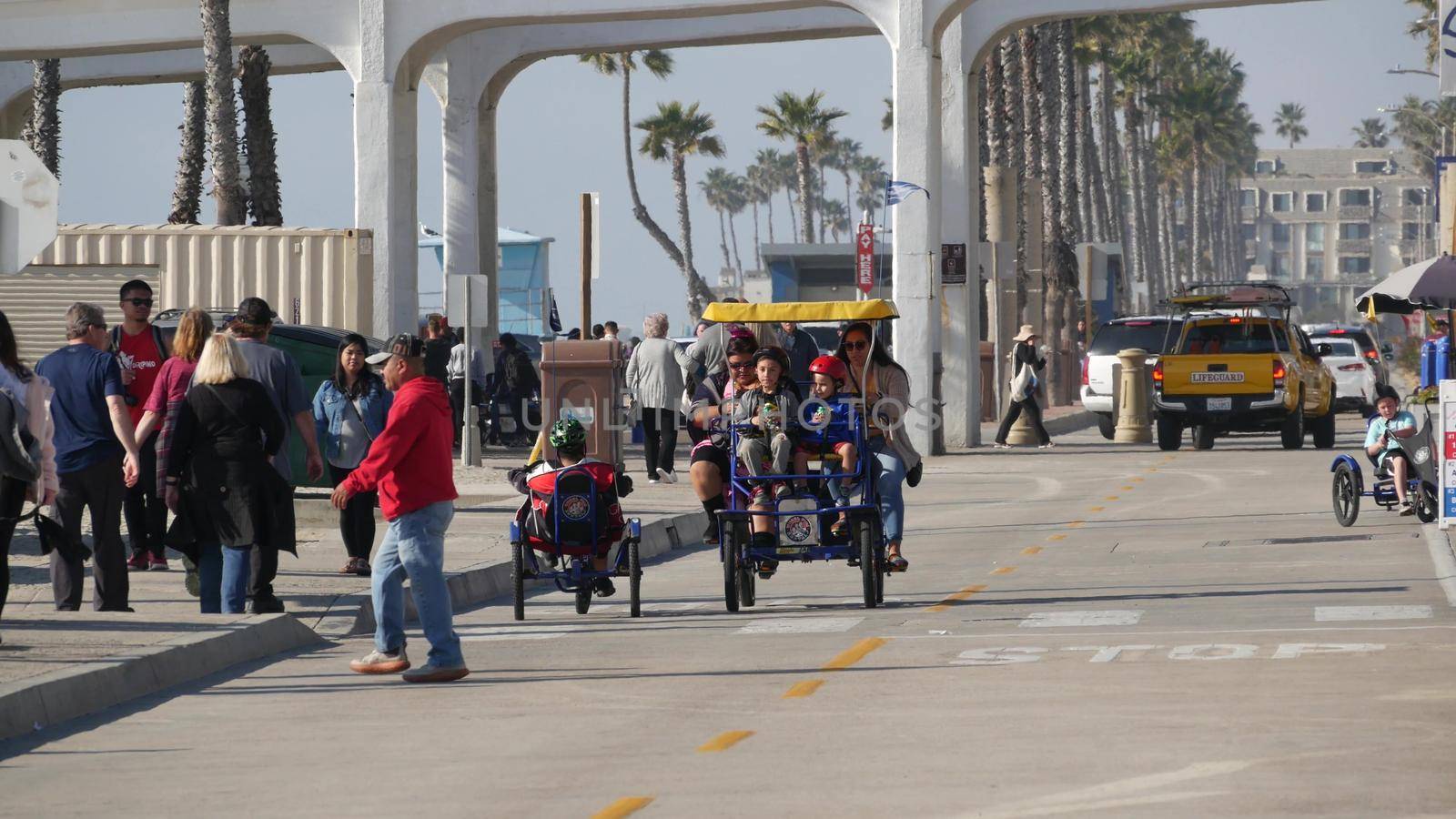 Oceanside, California USA - 8 Feb 2020: People walking on waterfront promenade, beachfront boardwalk. Vacations ocean beach resort near Los Angeles. Family riding surrey 4 wheel double bench bike.