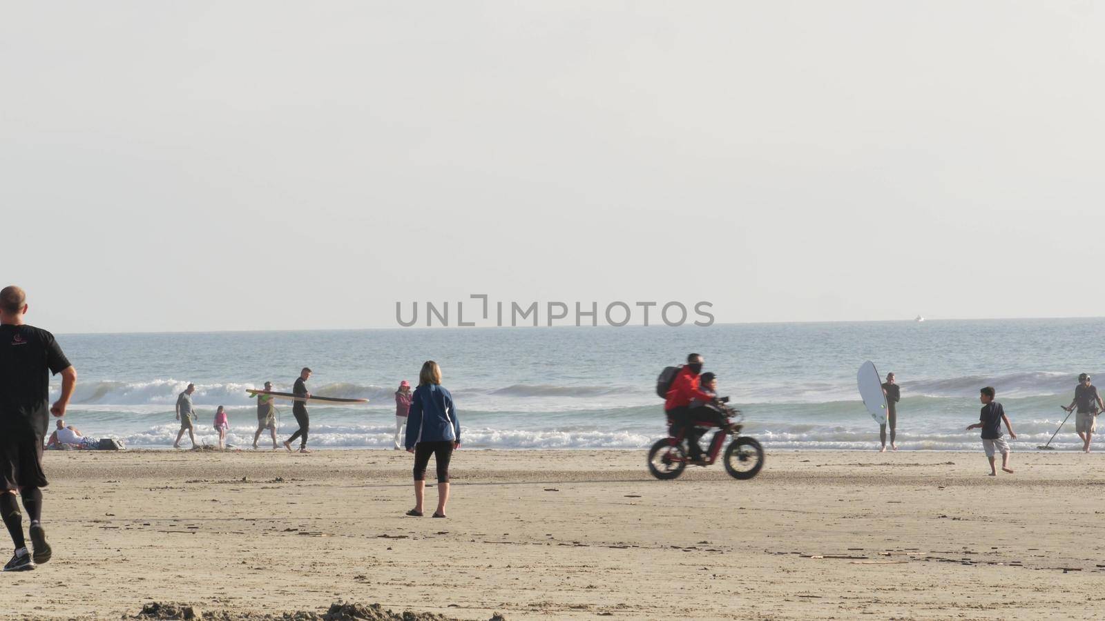 Oceanside, California USA - 8 Feb 2020: People walking on ocean beach, waterfront vacations resort. Surfer men with surfboard going surfing in sea waves. Family riding bikes. Man with metal detector.