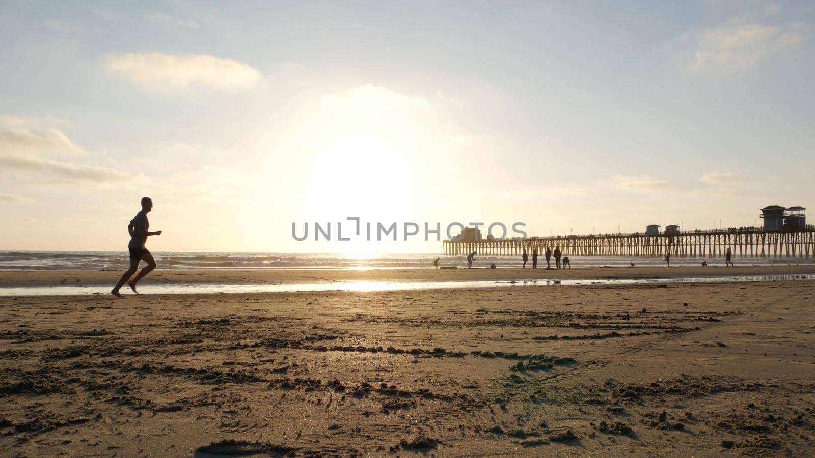 Oceanside, California USA - 8 Feb 2020: People walking on ocean beach, waterfront vacations resort. Contrast silhouette, man running, sand near sea waves. Young guy training in sunset light near pier.