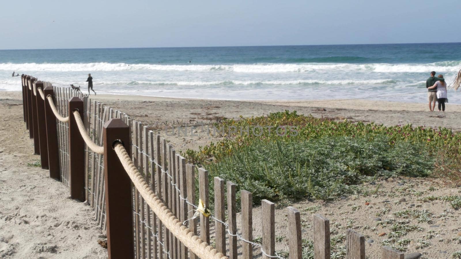 Encinitas, California USA - 23 Feb 2020: Pacific coast, people walking on ocean beach by sea water waves. Coastal access with picket fence on sandy shore near San Diego and Los Angeles. Couple and dog