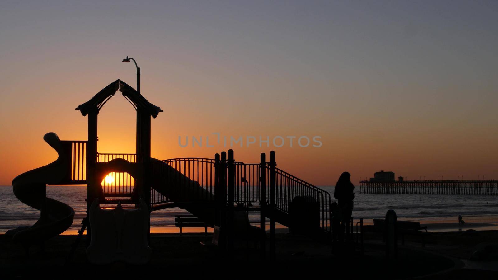 Oceanside, California USA - 11 Feb 2020: Silhouette of waterfront playground near pier. Children playing with parents, sunset ocean beach. Family with kids, beachfront recreation area, pacific coast.