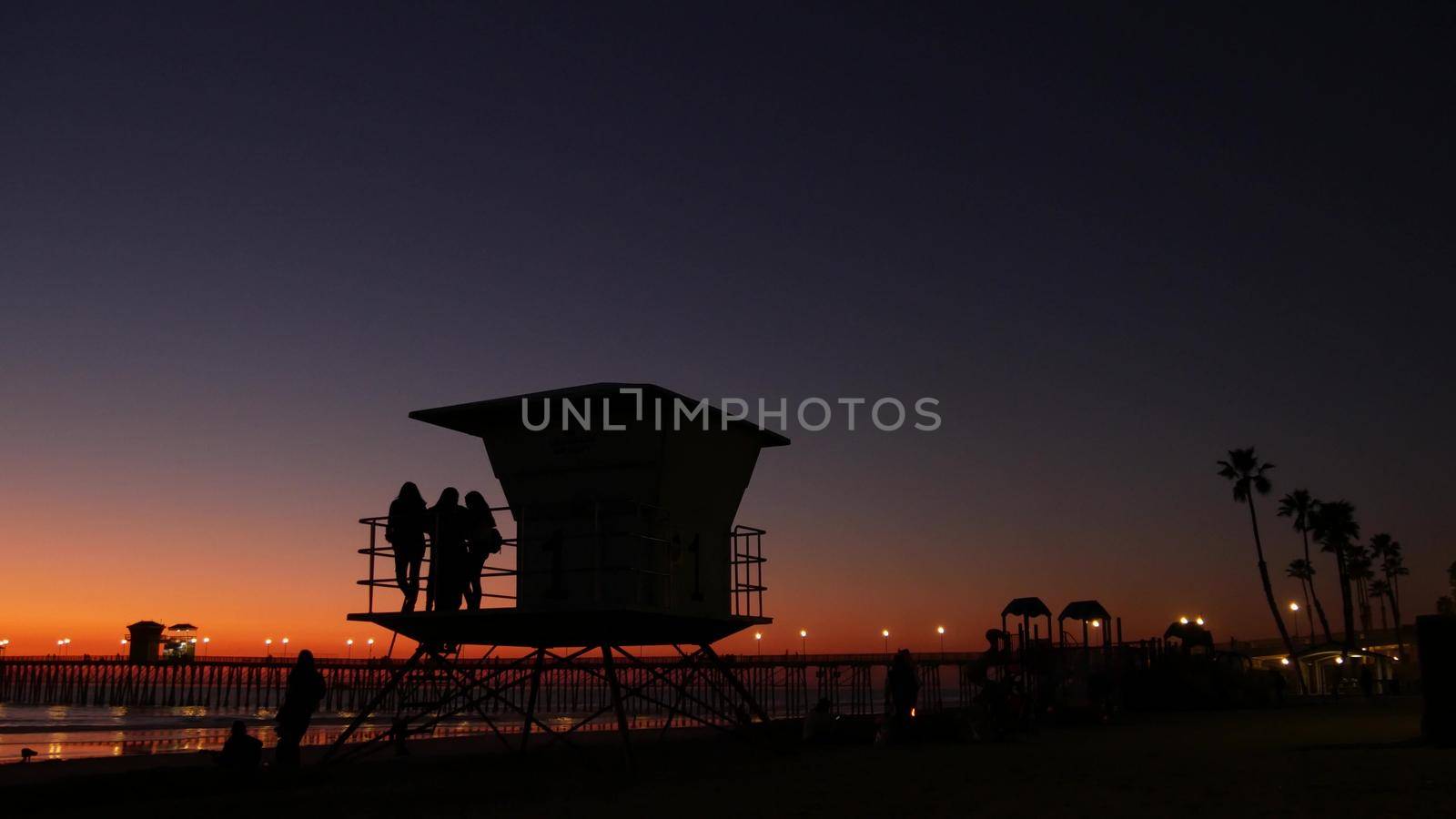 Oceanside, California USA - 8 Feb 2020: Young teen girls near lifeguard tower, friends on pacific ocean beach, sunset dusk. Unrecognizable teenagers, people and twilight gradient purple violet sky.