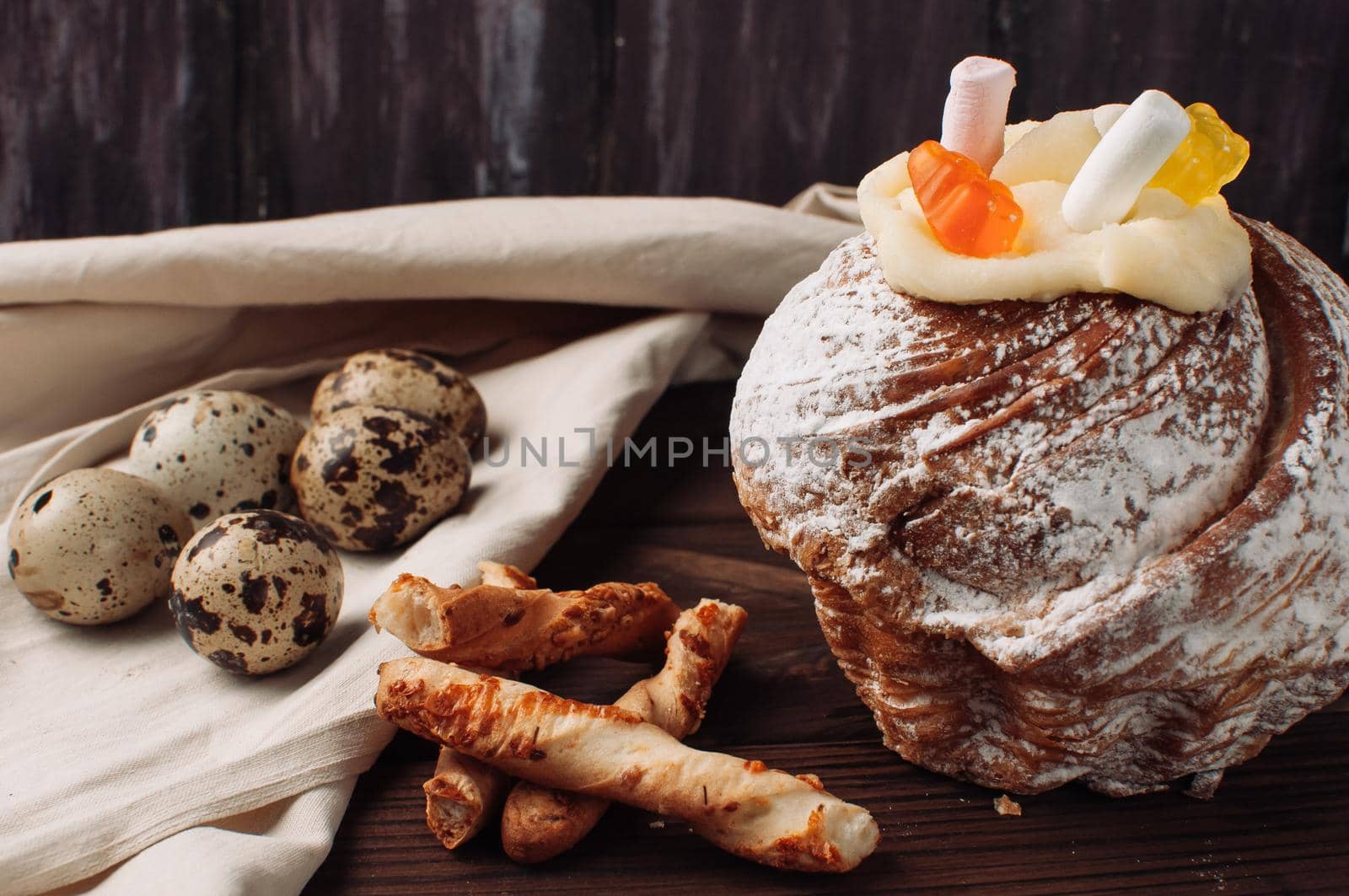 Stylish Easter cake with marshmallows and jelly bears on a dark rustic wooden background, next to it are quail eggs on a linen tablecloth and Italian grissini. Seasonal greetings for Easter.