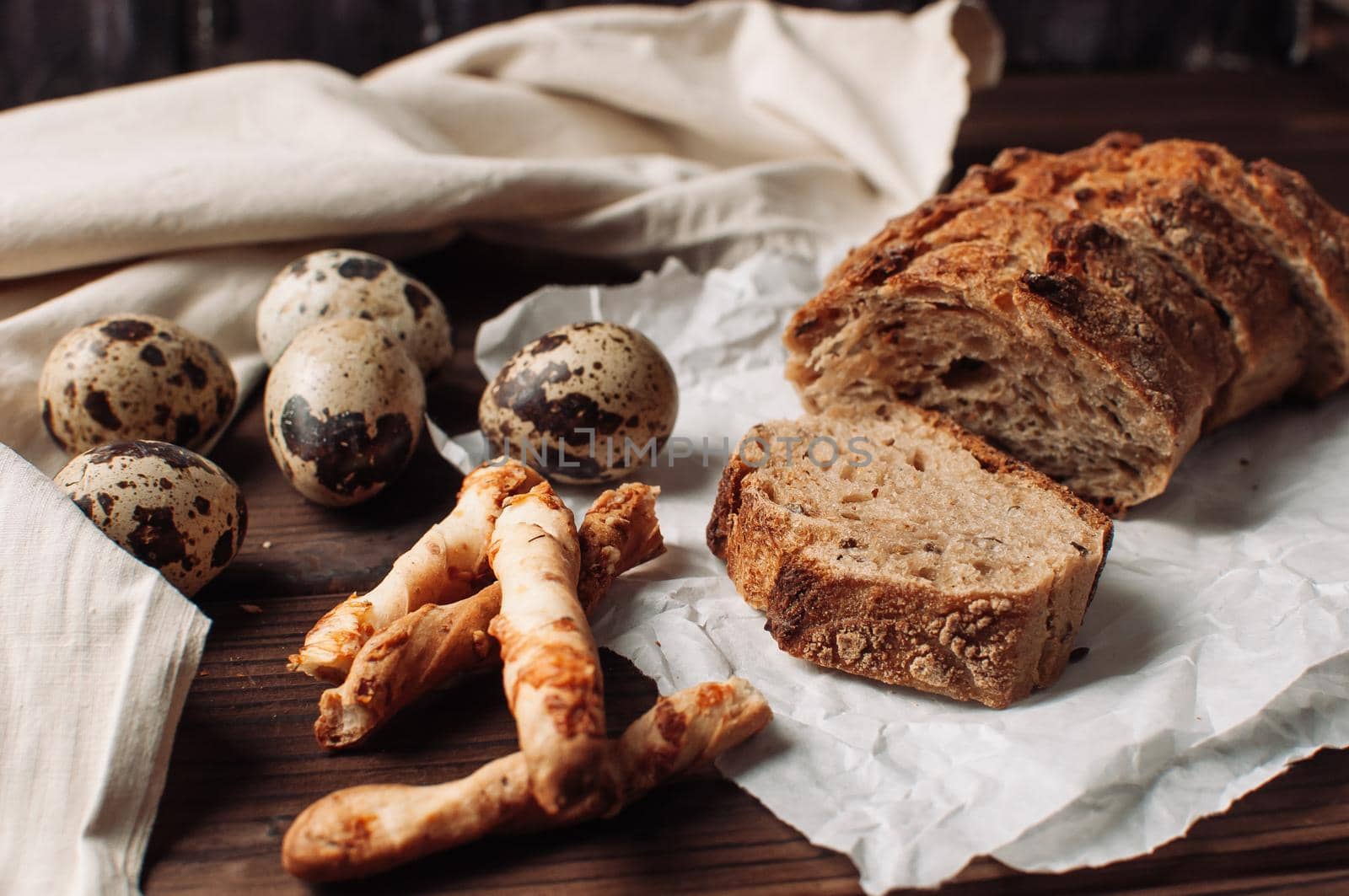 set dark yeast-free buckwheat bread in a cut lies on parchment, next to quail eggs and Italian grissini on a linen tablecloth on a wooden table in a rustic style. Breakfast cooking concept.
