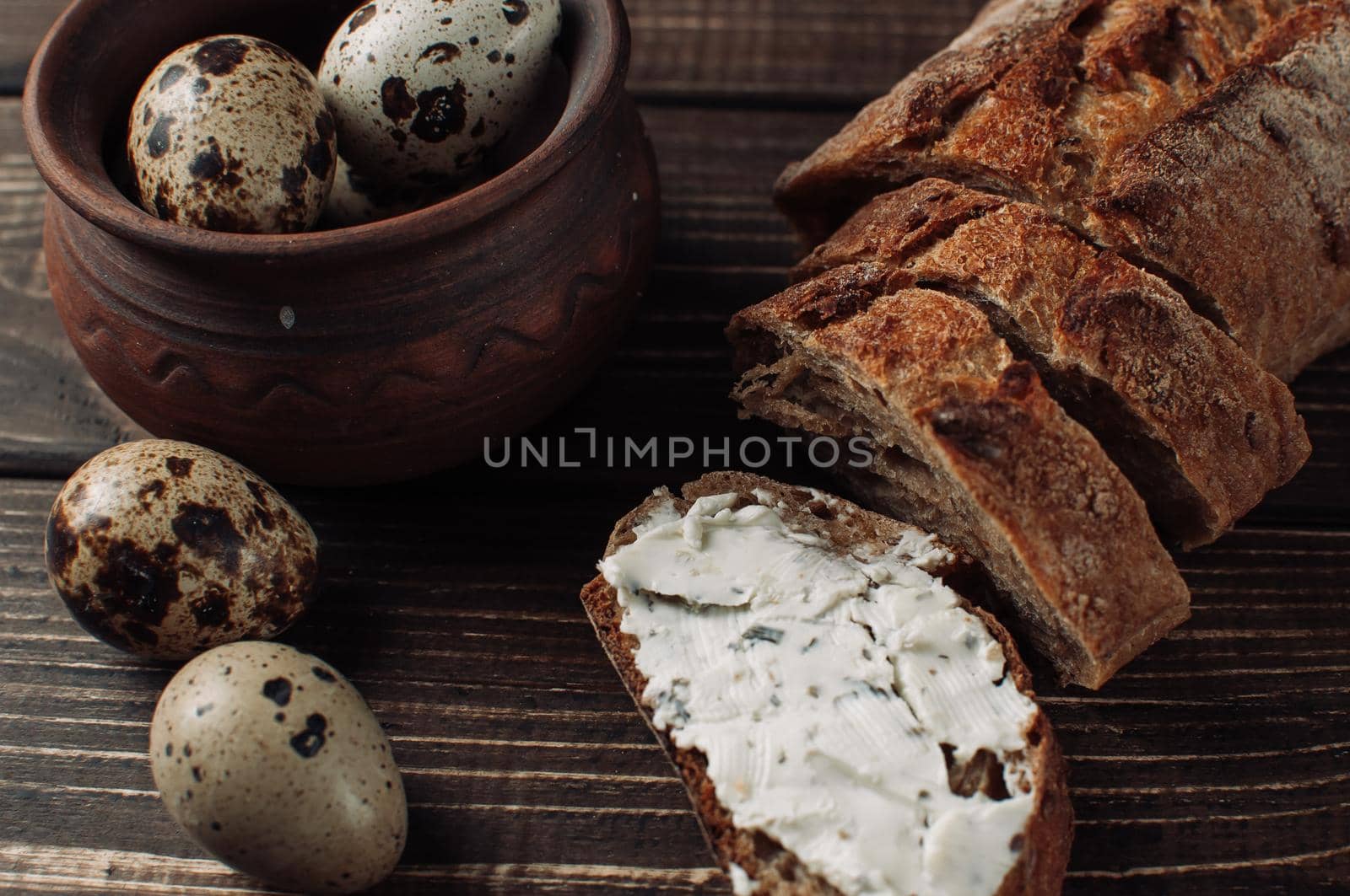 dark buckwheat bread is spread with cottage cheese with herbs in a cut on a wooden table near quail eggs in a clay plate in a rustic style. Snack and breakfast concept.
