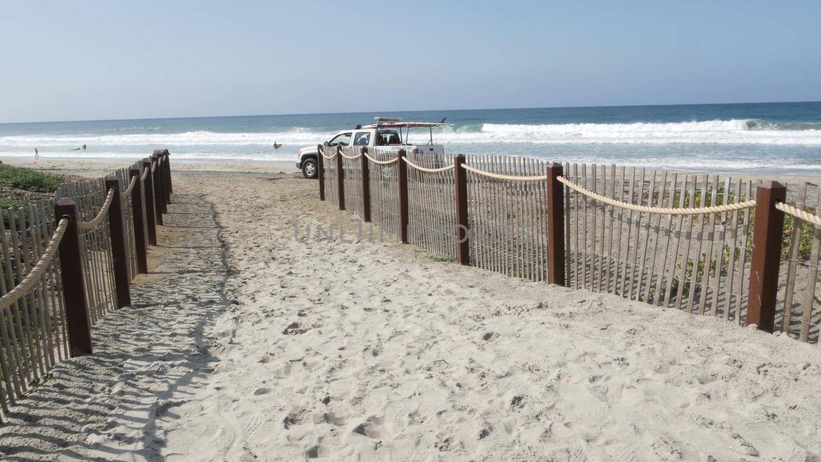Encinitas, California USA - 23 Feb 2020: Pacific coast, people walking on ocean beach by sea water waves. Coastal access with picket fence on sandy shore near San Diego and Los Angeles. Lifeguard car.