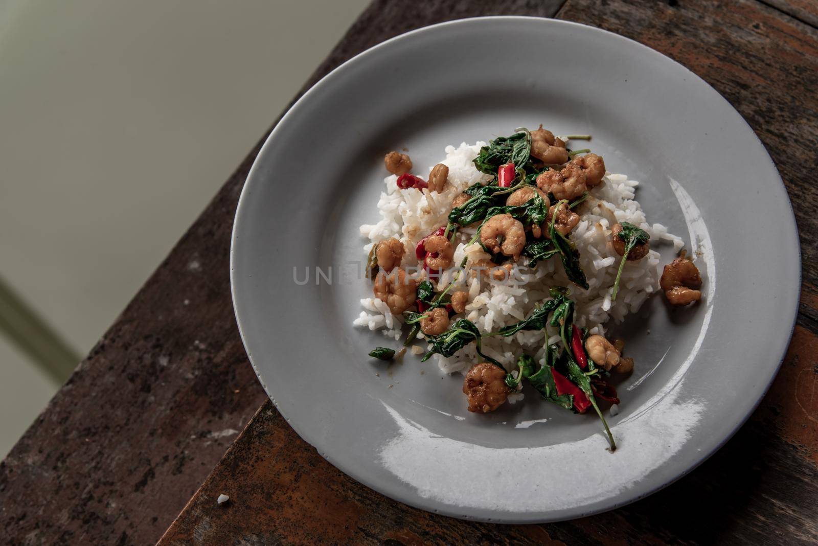 Rice topped with Stir fried thai basil with dwarf prawn served in white dish on wooden table, A world renowned delicacy from Thailand, Selective focus.