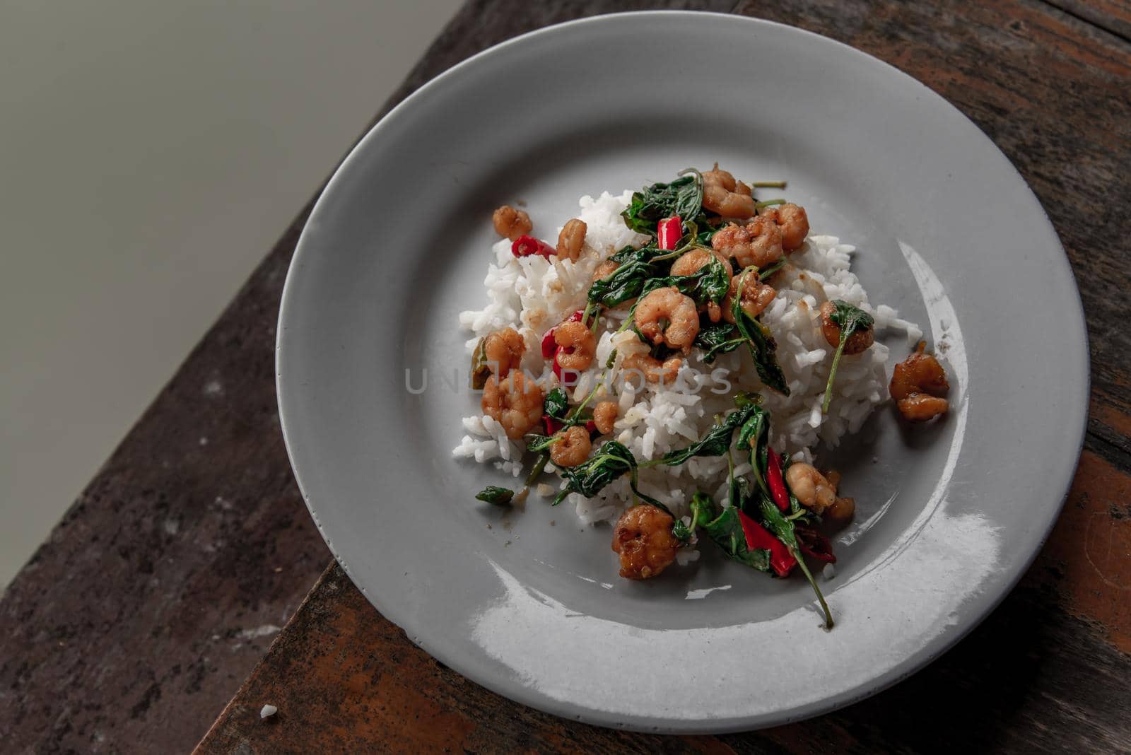 Rice topped with Stir fried thai basil with dwarf prawn served in white dish on wooden table, A world renowned delicacy from Thailand.