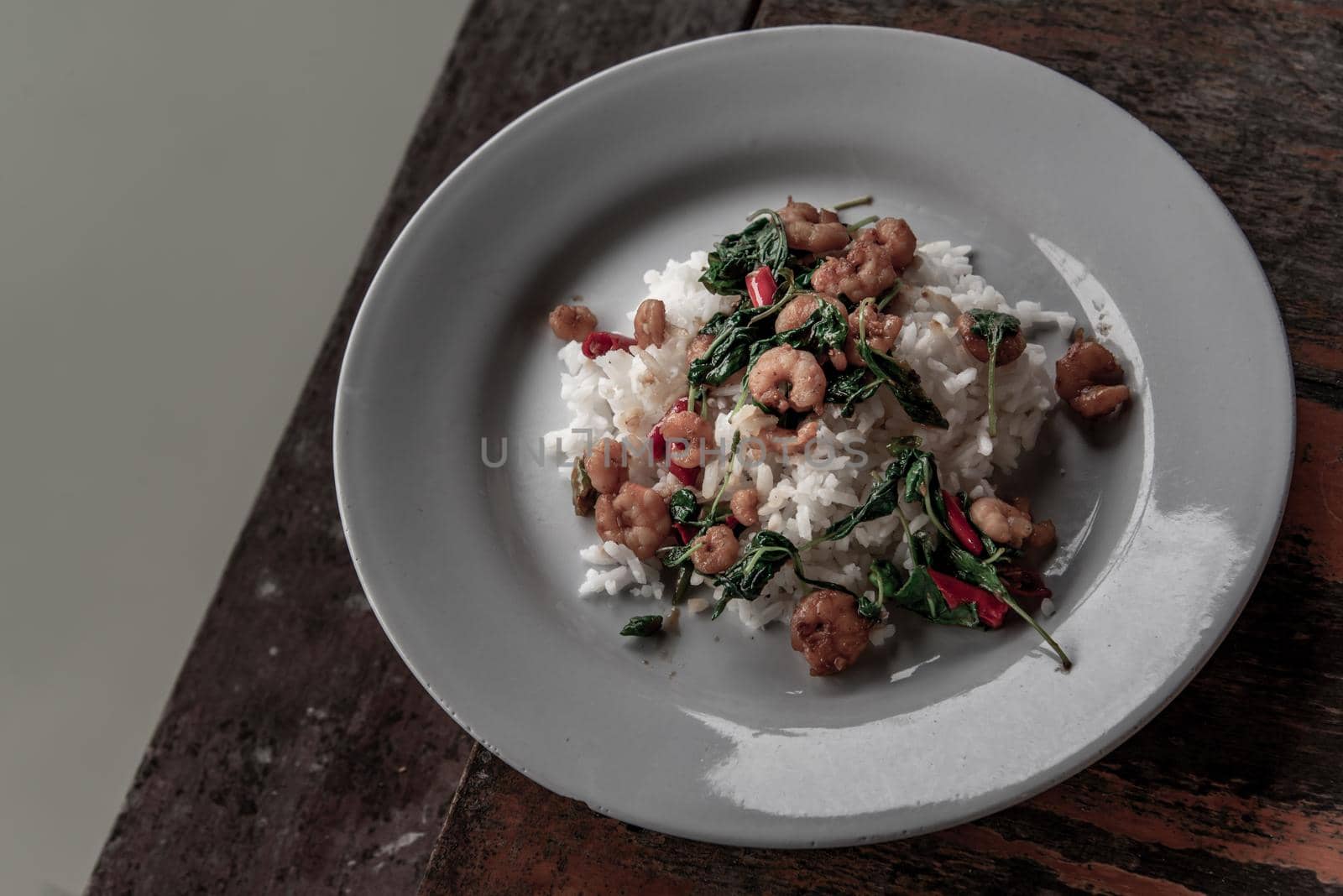 Rice topped with Stir fried thai basil with dwarf prawn served in white dish on wooden table, A world renowned delicacy from Thailand.