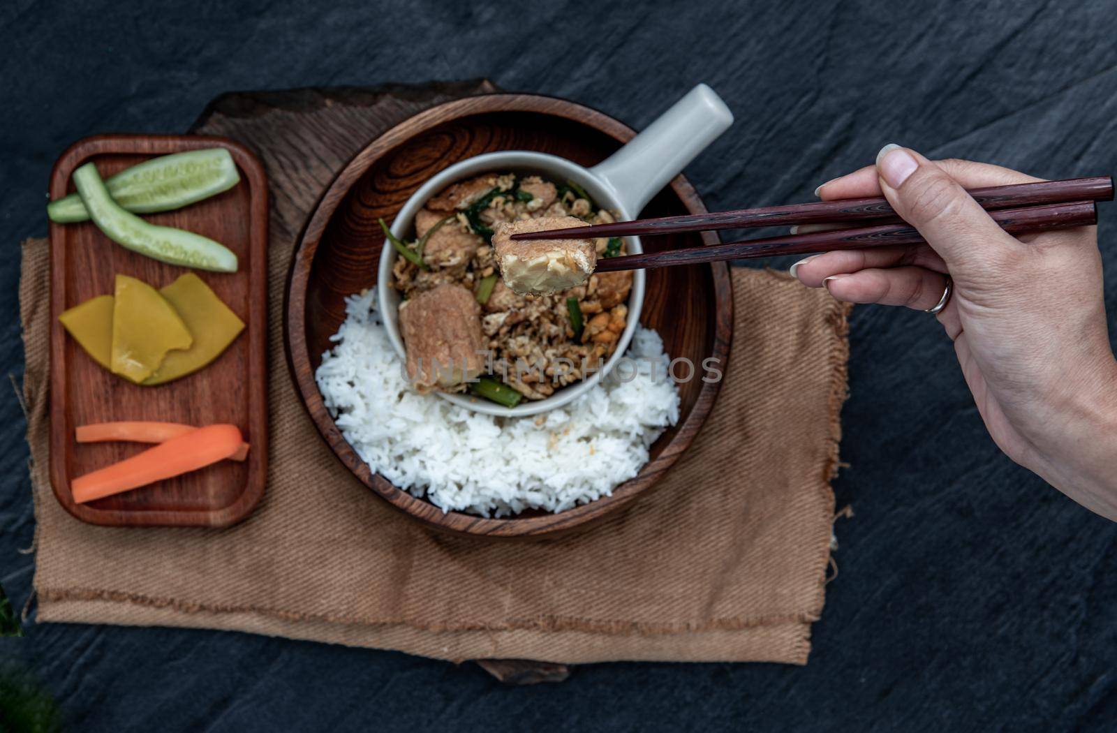 Thai food, The hand is using chopsticks to pick up  Stir fried egg tofu with spring onion with Herbs vegetables served with steamed rice in wooden bowl.