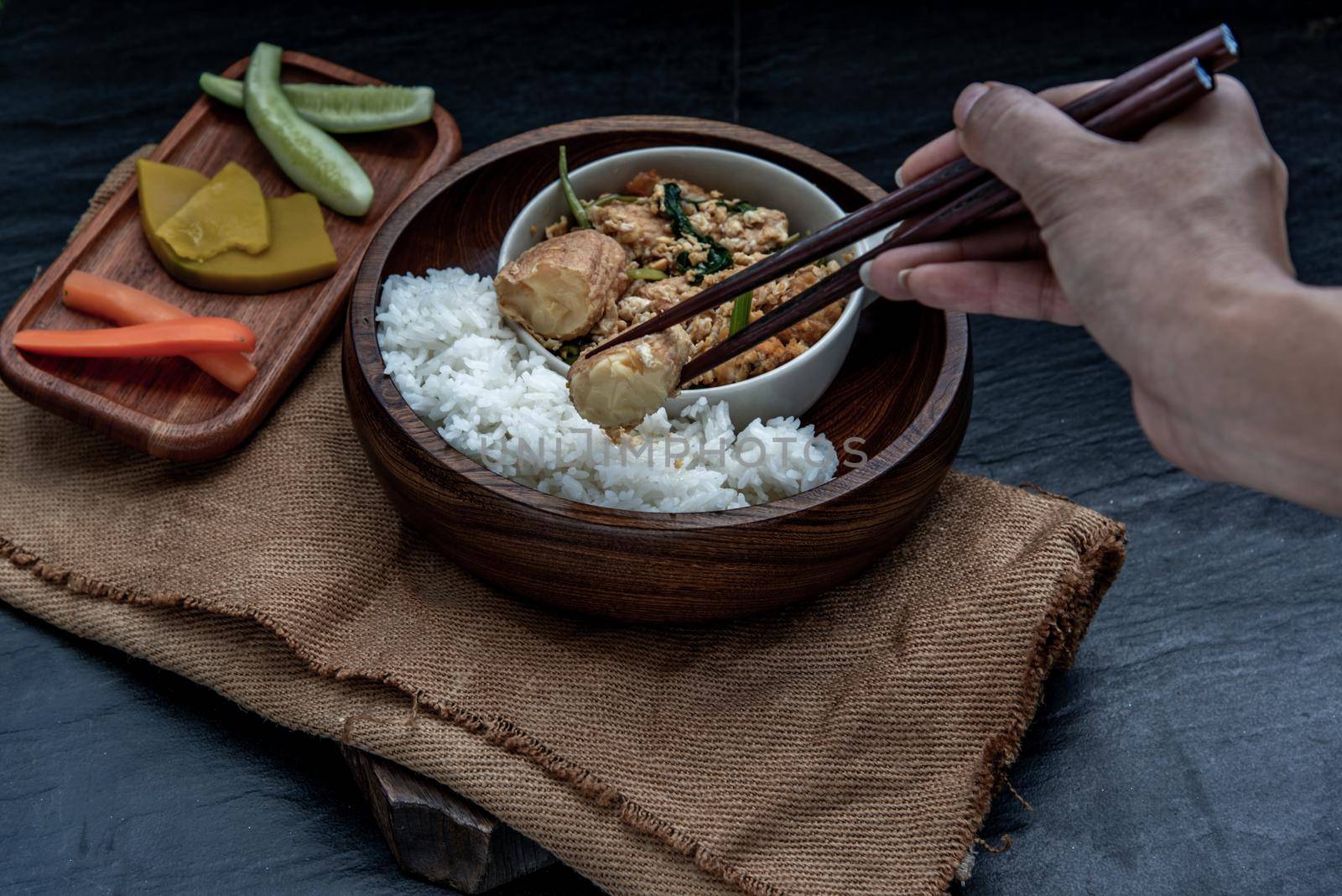 Thai food, The hand is using chopsticks to pick up  Stir fried egg tofu with spring onion with Herbs vegetables served with steamed rice in wooden bowl.