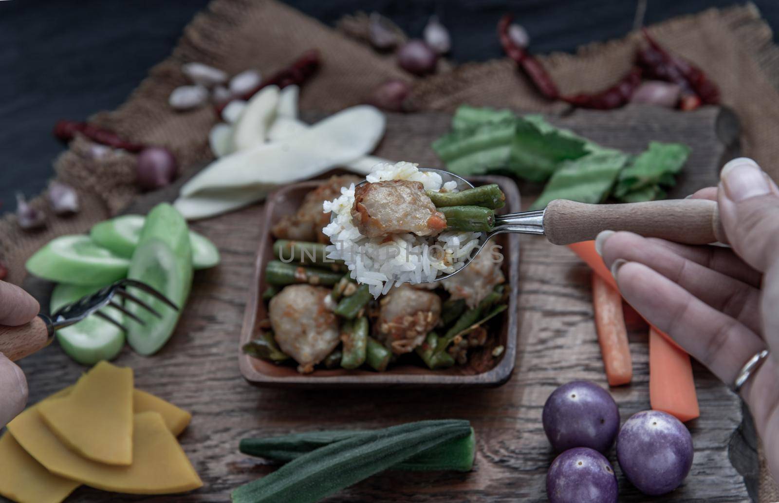 Thai food, The hand is using a spoon to scoop Stir Fried fish ball with Red Curry Paste and long bean with the ingredient and fresh vegetables on wood background.