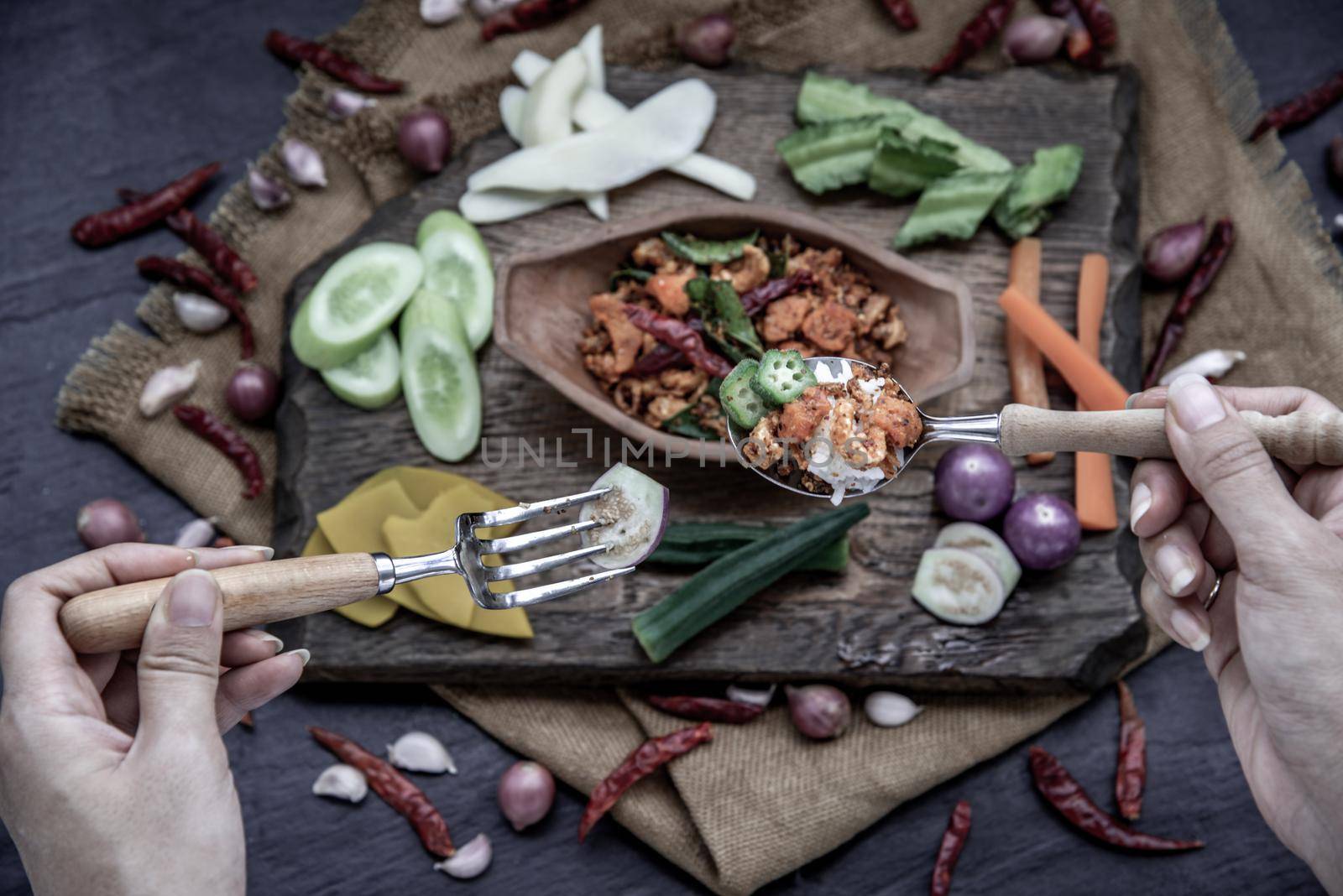 The hand is using a spoon to scoop Pork Crackling Chili Paste with fried kaffir lime leaves with the ingredient and fresh vegetables on wood background, view from above.