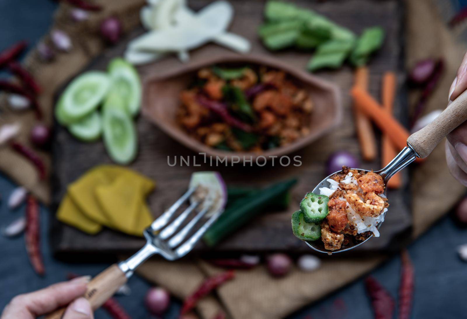 The hand is using a spoon to scoop Pork Crackling Chili Paste with fried kaffir lime leaves with the ingredient and fresh vegetables on wood background, view from above.