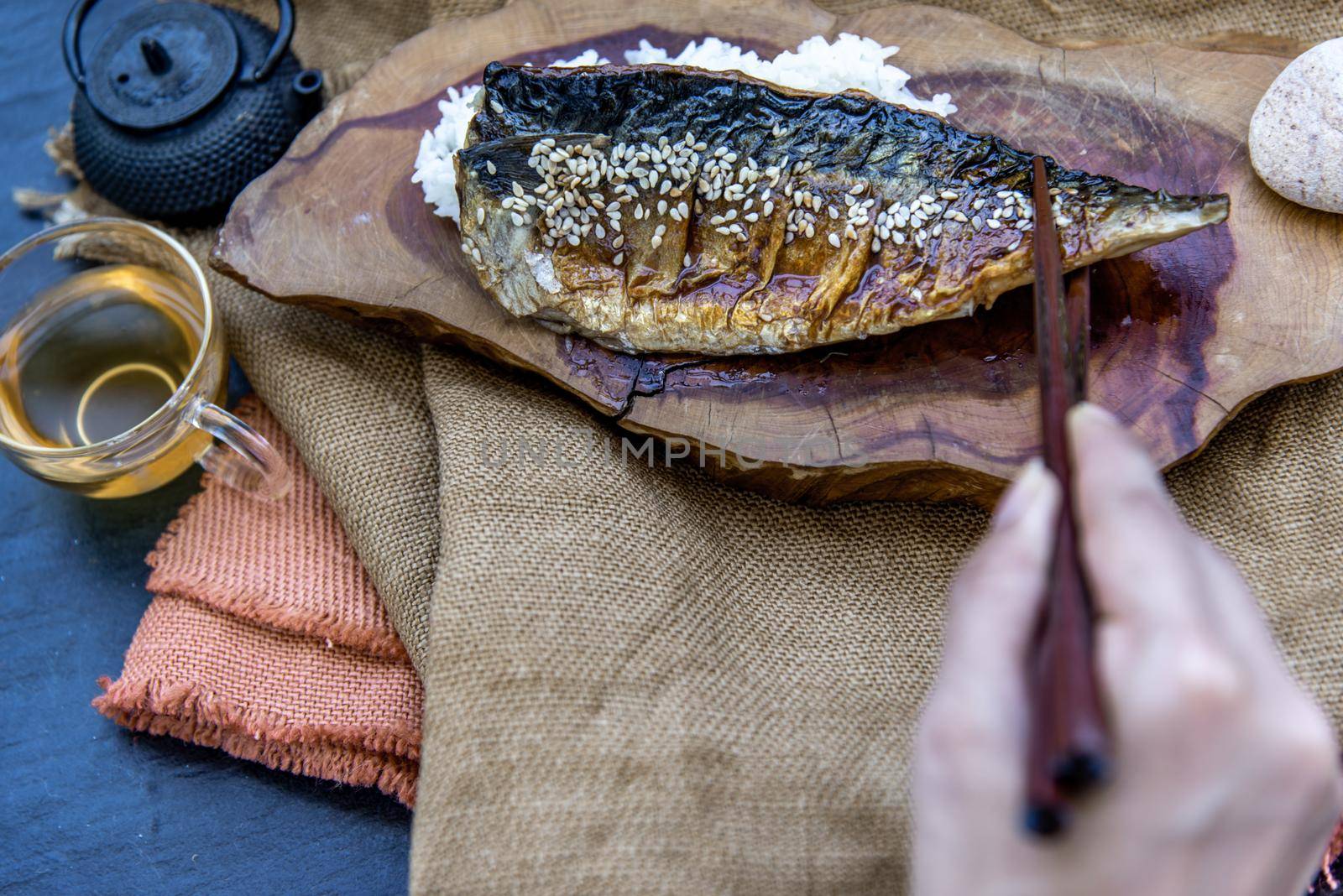 The hand is using chopsticks to pick up saba fish grilled on rice with teriyaki sauce on a wooden background served with hot tea, Japanese style.