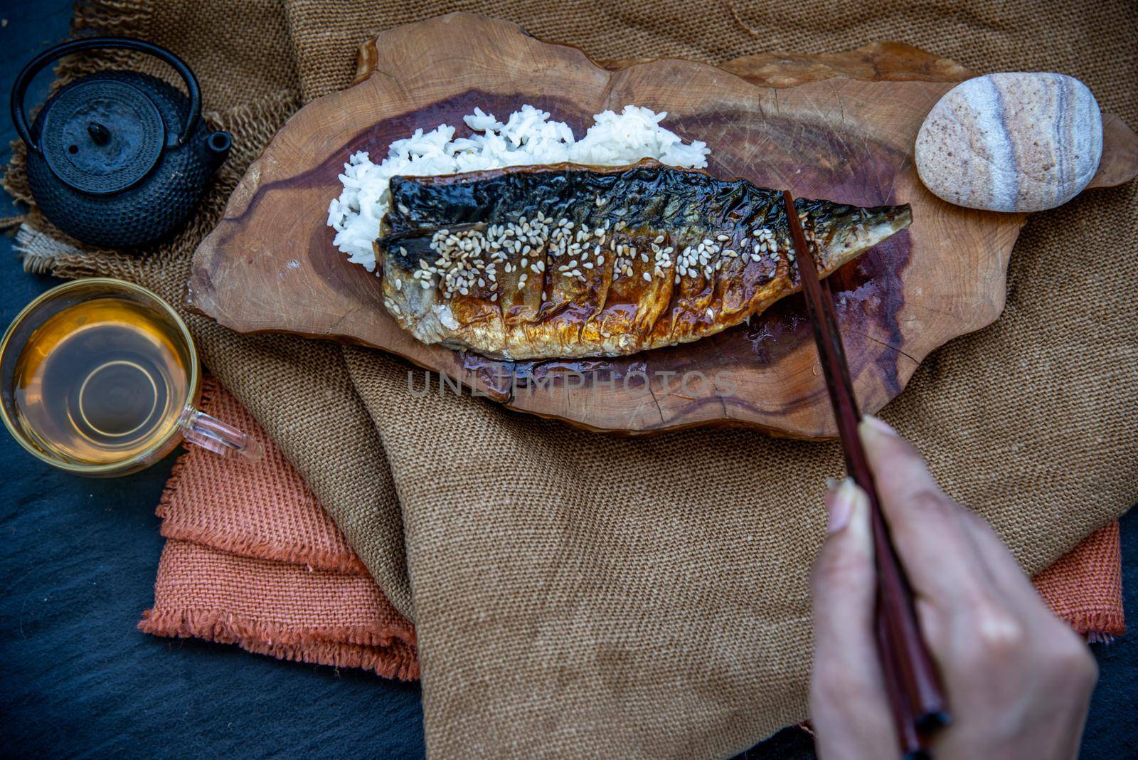 The hand is using chopsticks to pick up saba fish grilled on rice with teriyaki sauce on a wooden background served with hot tea, Japanese style.