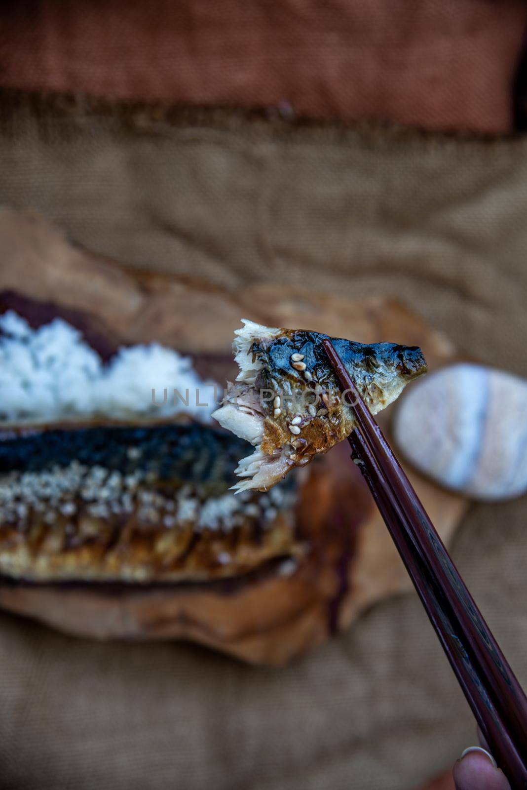 The hand is using chopsticks to pick up saba fish grilled on rice with teriyaki sauce on a sackcloth background, Japanese style.