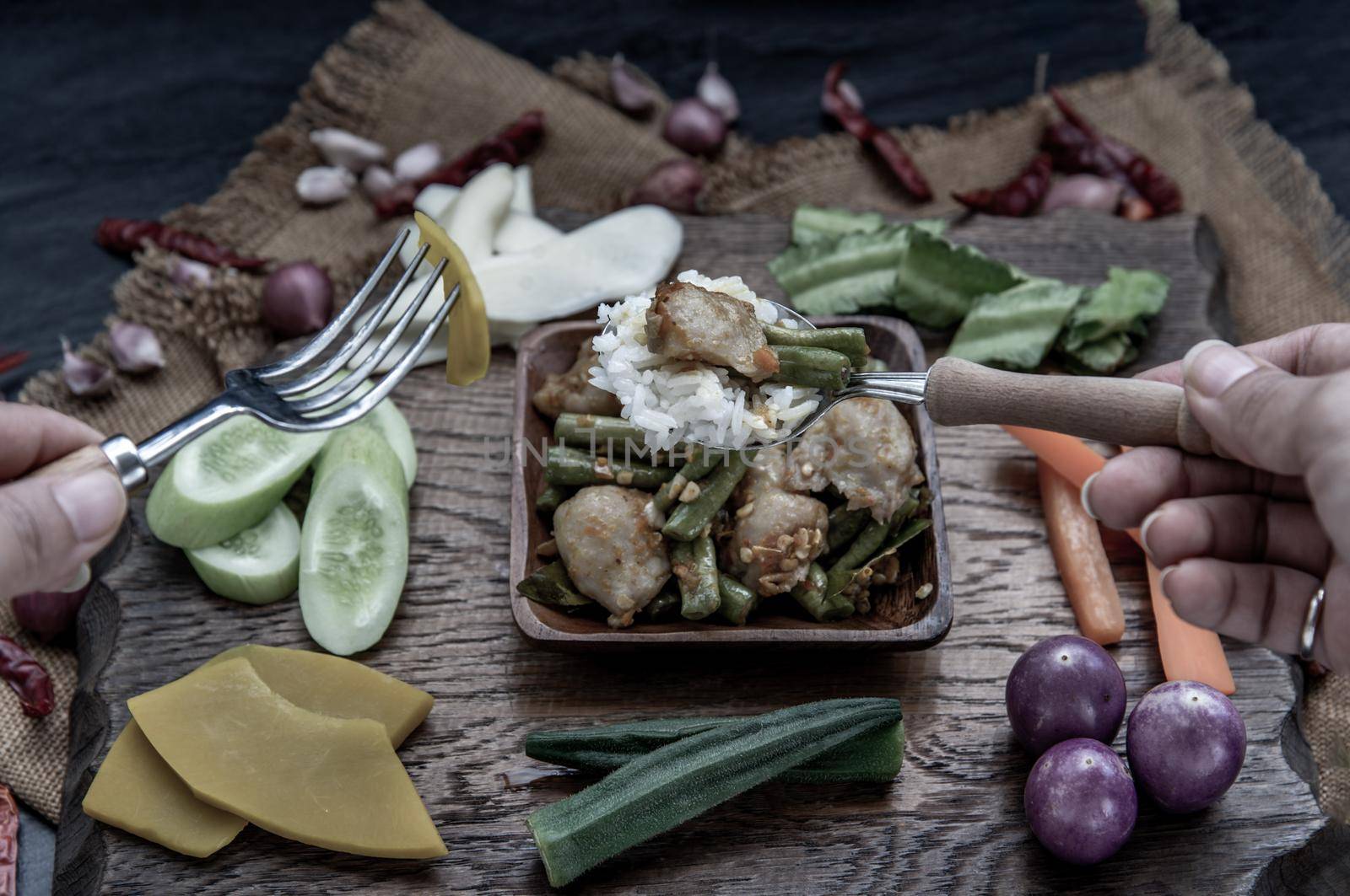 Thai food, The hand is using a spoon to scoop Stir Fried fish ball with Red Curry Paste and long bean with the ingredient and fresh vegetables on wood background.