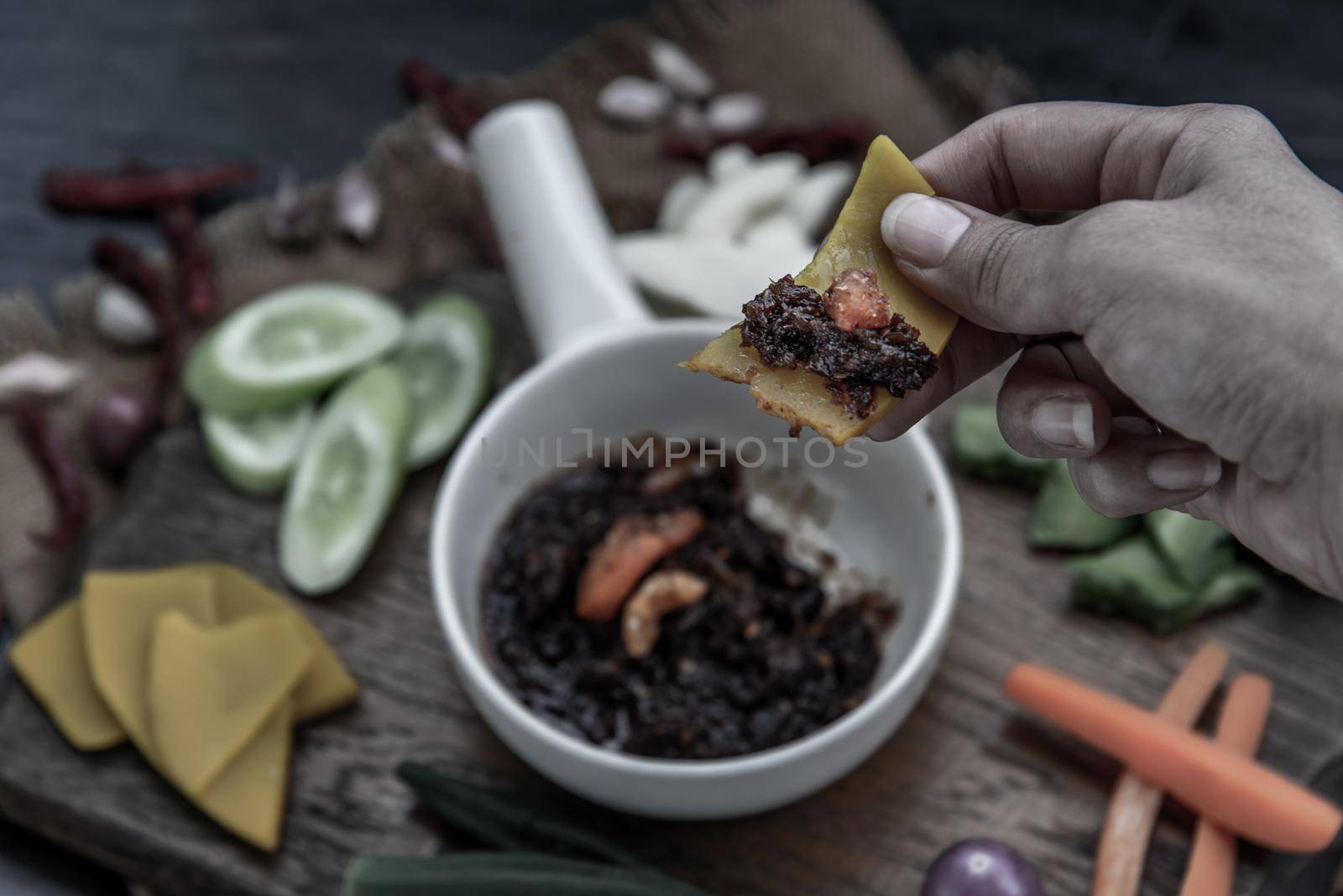 The hand is using pumpkin to scoop fried chili paste with shrimps (thai language nam prik pao) with the ingredient and fresh vegetables on wood background. Thai food.