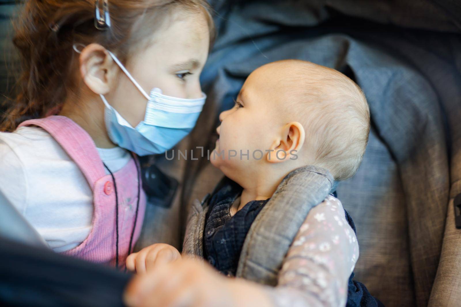 Portrait of adorable little girl wearing pink overalls and face mask next to her happy baby sister in stroller. Close-up of cute young children with blurry background. New normal after Covid-19