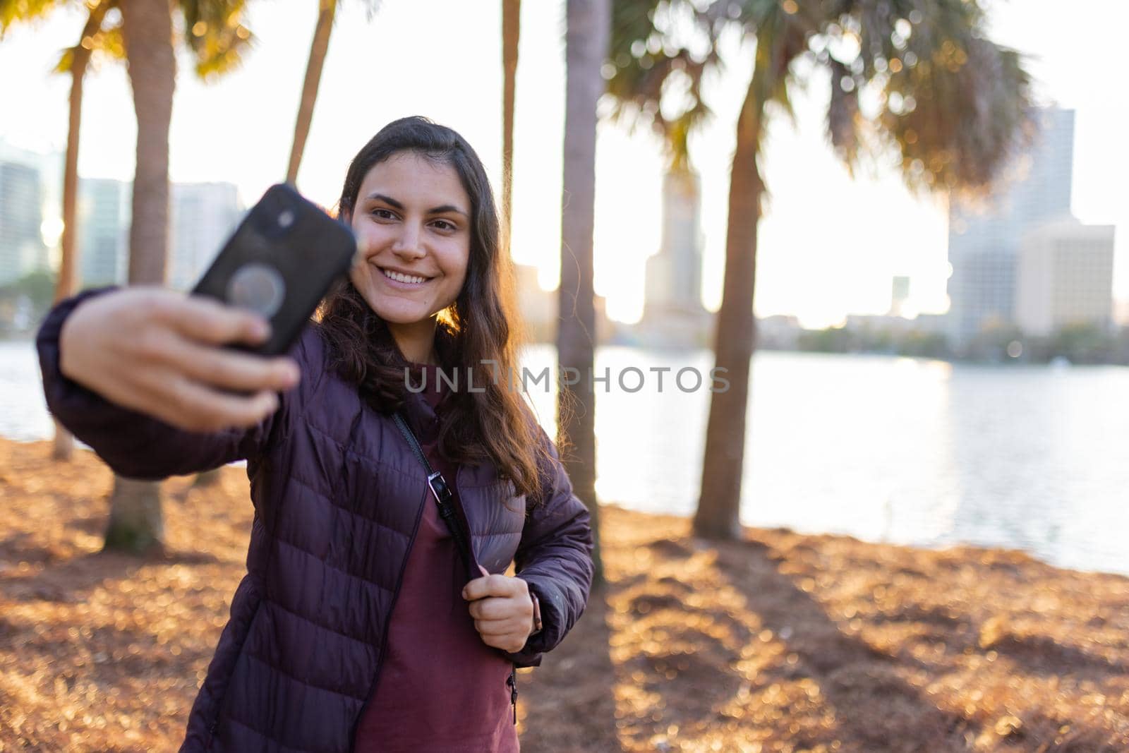 Beautiful view of smiling woman taking selfie with blurry palm trees, and bright sunset as background. Happy brunette woman in black jacket taking self-portrait near a lake. Adventurous day outdoors