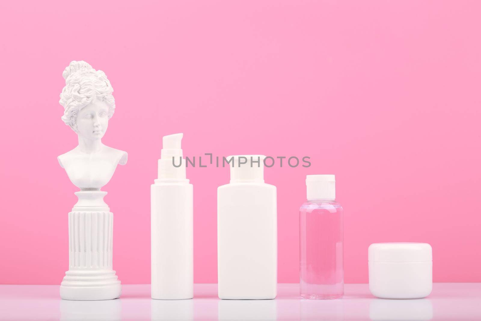 Set of beauty products for daily skin care on white table against bright pink background with white gypsum statue. Unbranded cosmetic bottles for daily skin routine
