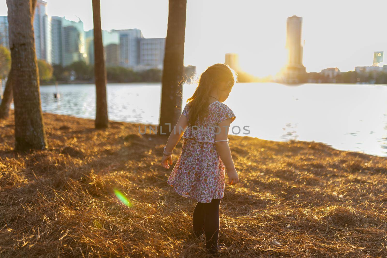 Warm view of cute little girl in colorful dress standing on grass with blurry lake and bright sunset as background. Sun shining on horizon behind young child in the evening. Kids playing outside