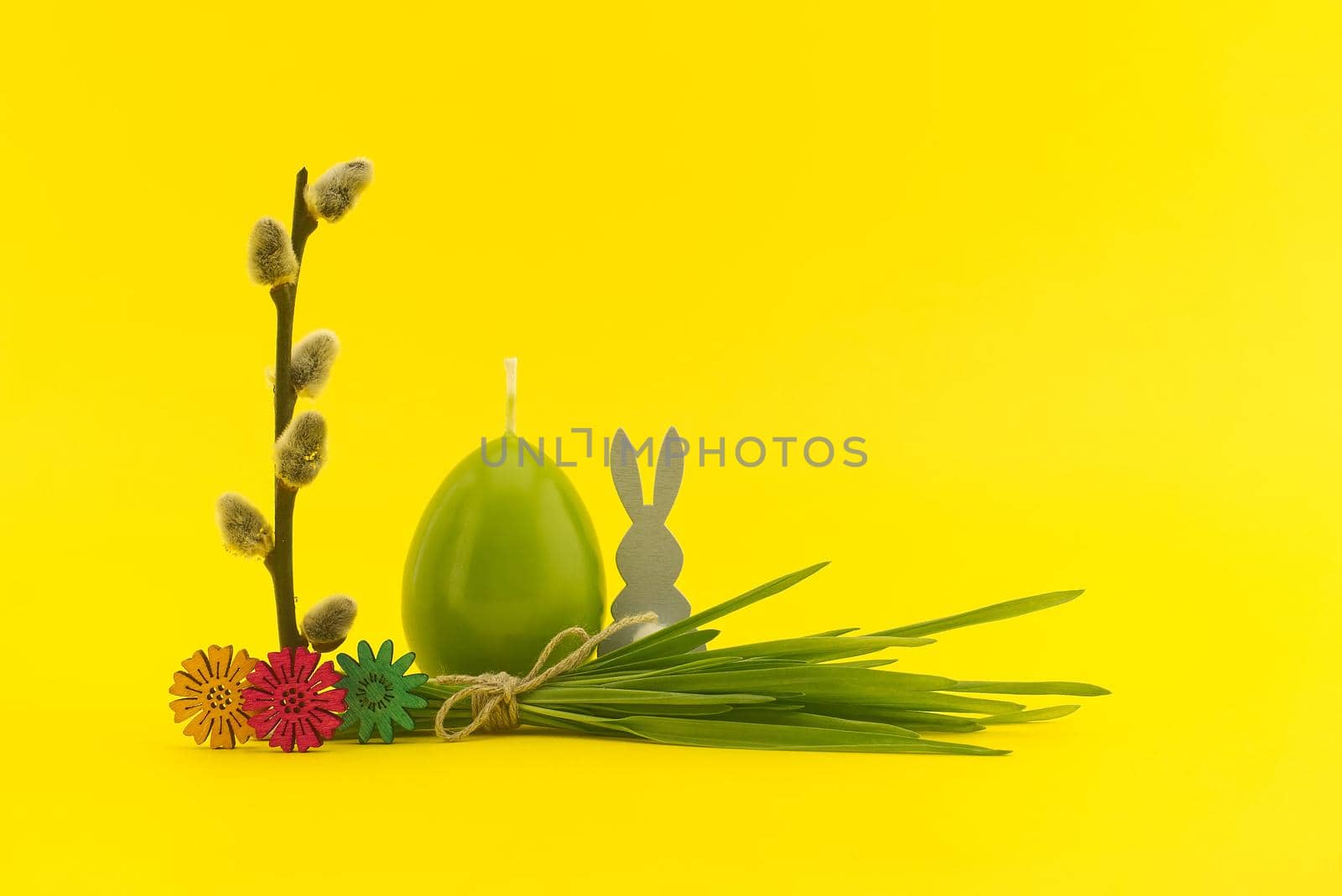 Creative Easter holiday or spring background with egg shaped candle, pussy willow branch and bundle of wheat seedlings tied with linen twine on yellow background with free copy space
