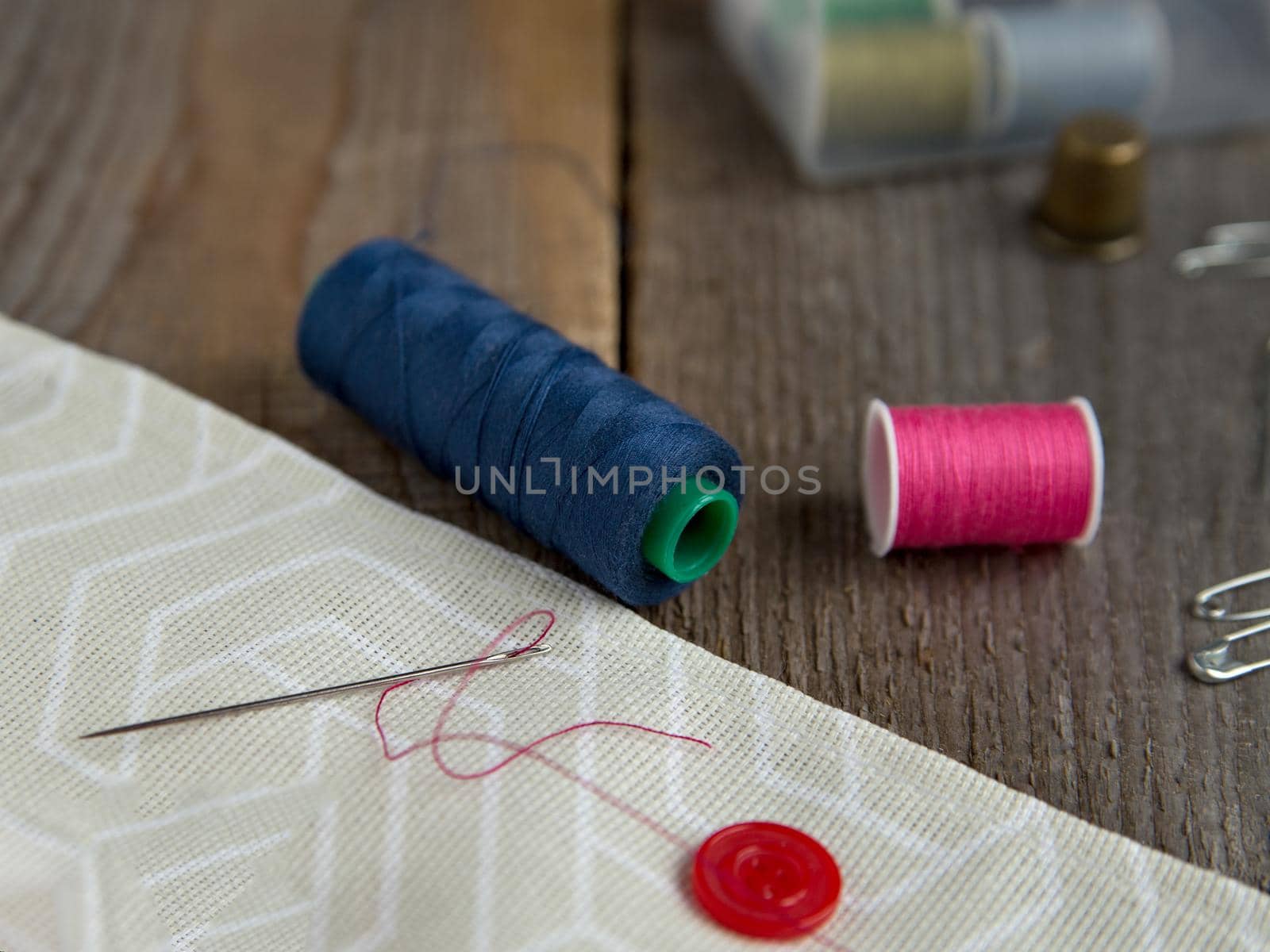 Coils with red and blue threads, needle with thread, gray fabric on brown wooden boards. Selective focus