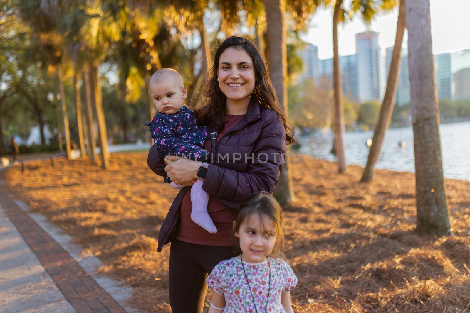 Beautiful view of happy mother and her adorable young daughters with trees and sunset sunlight as background. Portrait of little girl and smiling woman holding baby outside. Lovely family outdoors