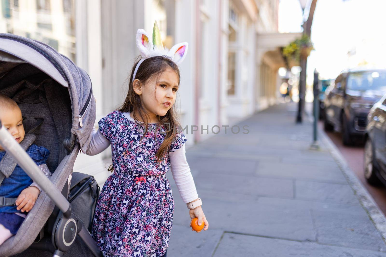 Adorable view of little girl wearing unicorn headband in the street next to her baby sister. Portrait of cute young child and gray stroller on the sidewalk. Lovely kids outdoors