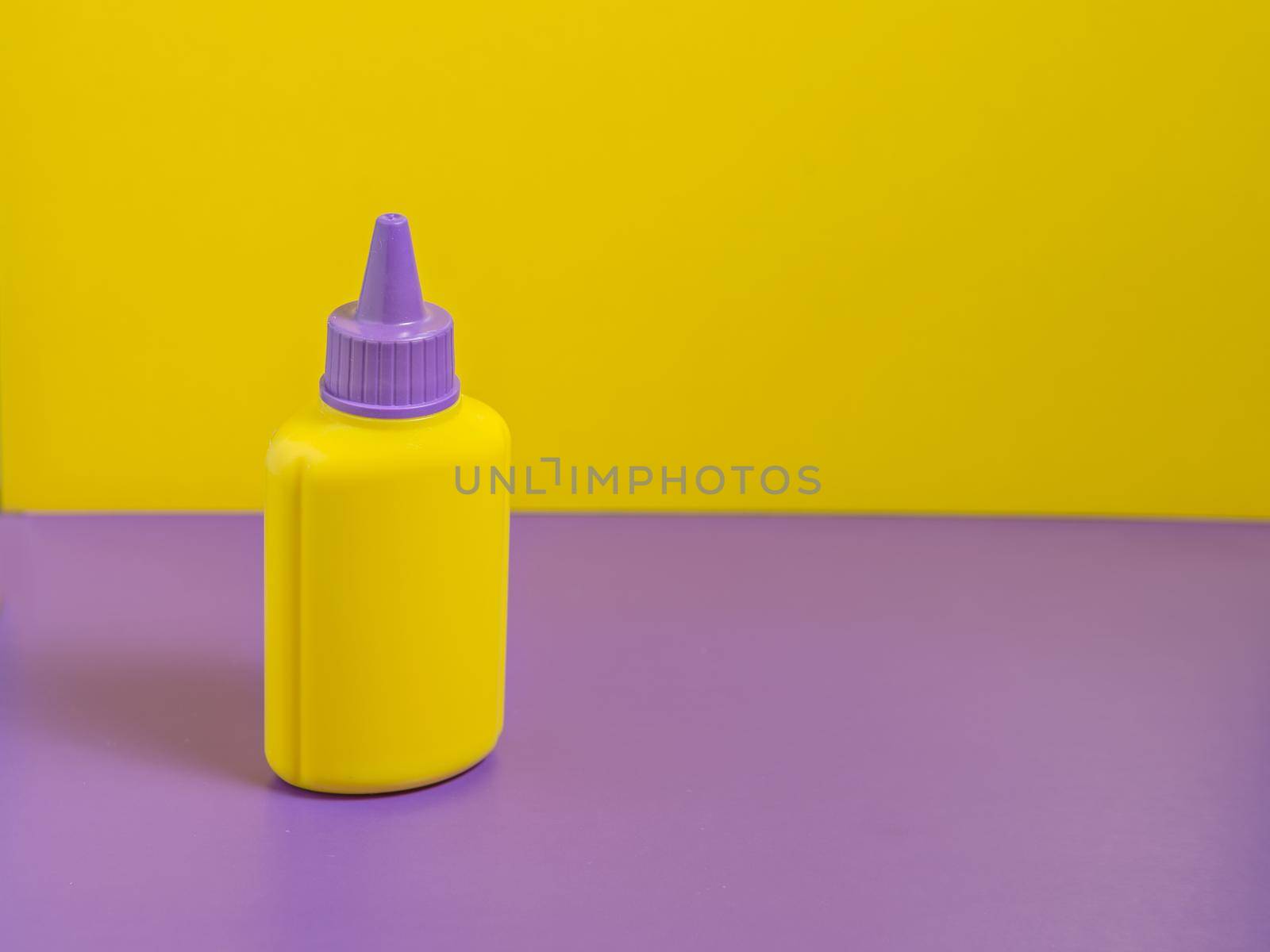 Yellow bottle with purple cap stands on purple table with yellow background.