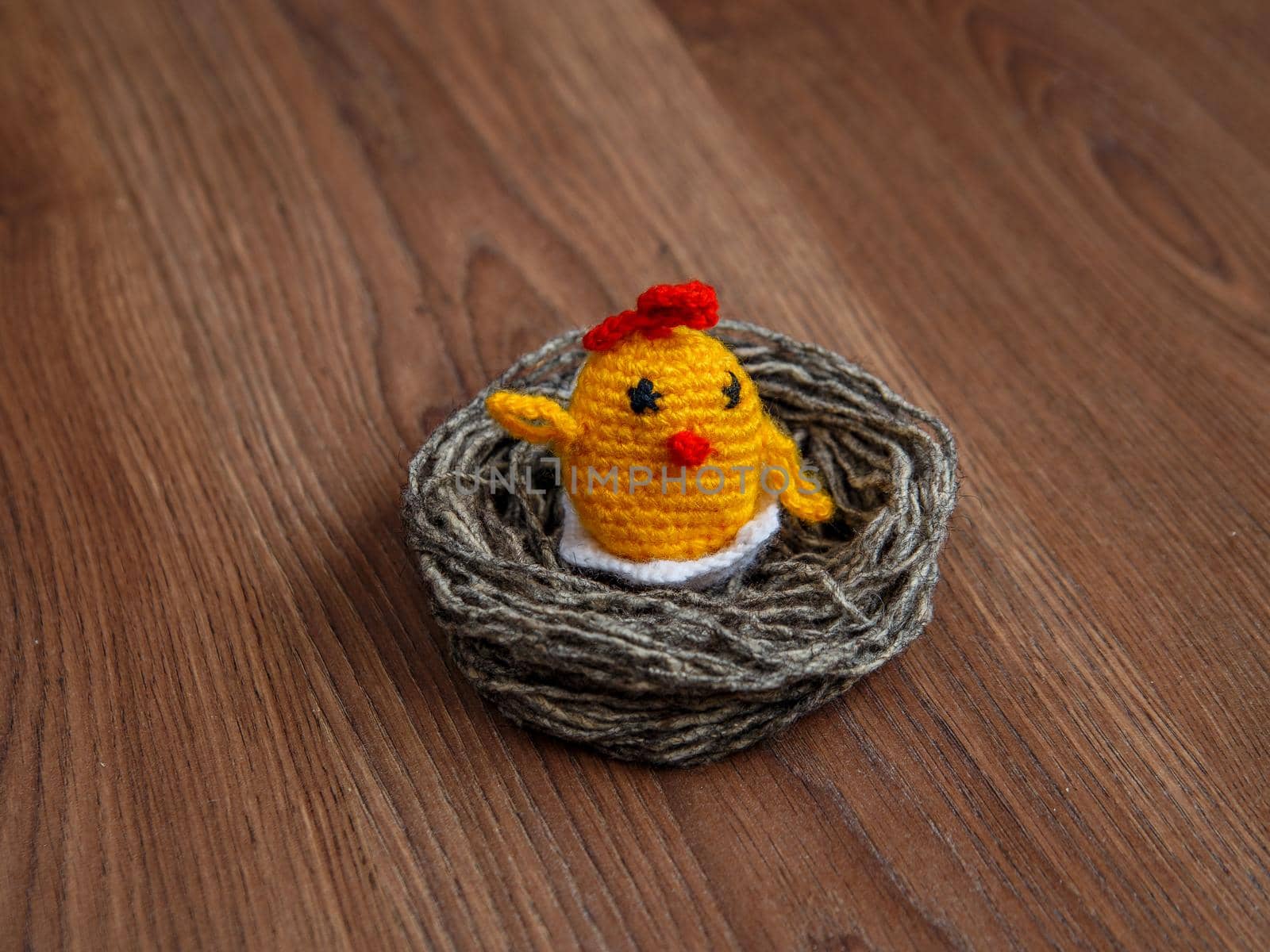 Yellow toy chicken in nest of gray woolen threads on brown surface.