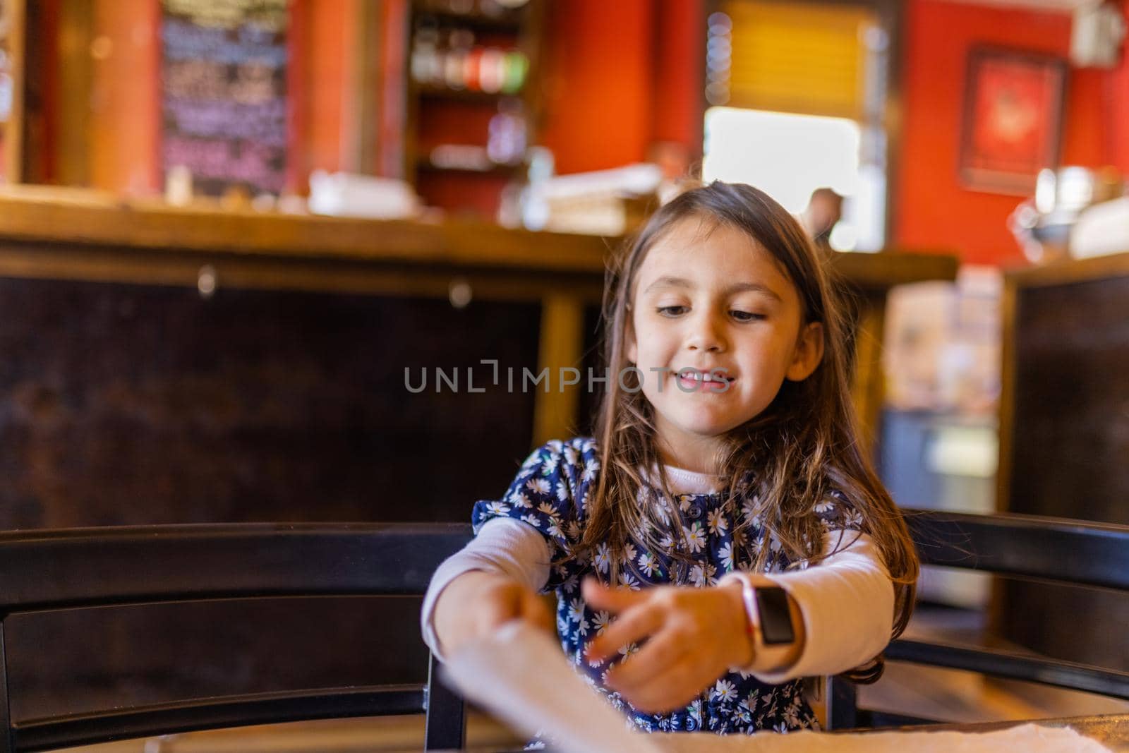 Adorable view of curious little girl in flowered dress adorably holding white napkin in restaurant. Cute young child sitting at a table playing with blurry background. Kids interacting with food