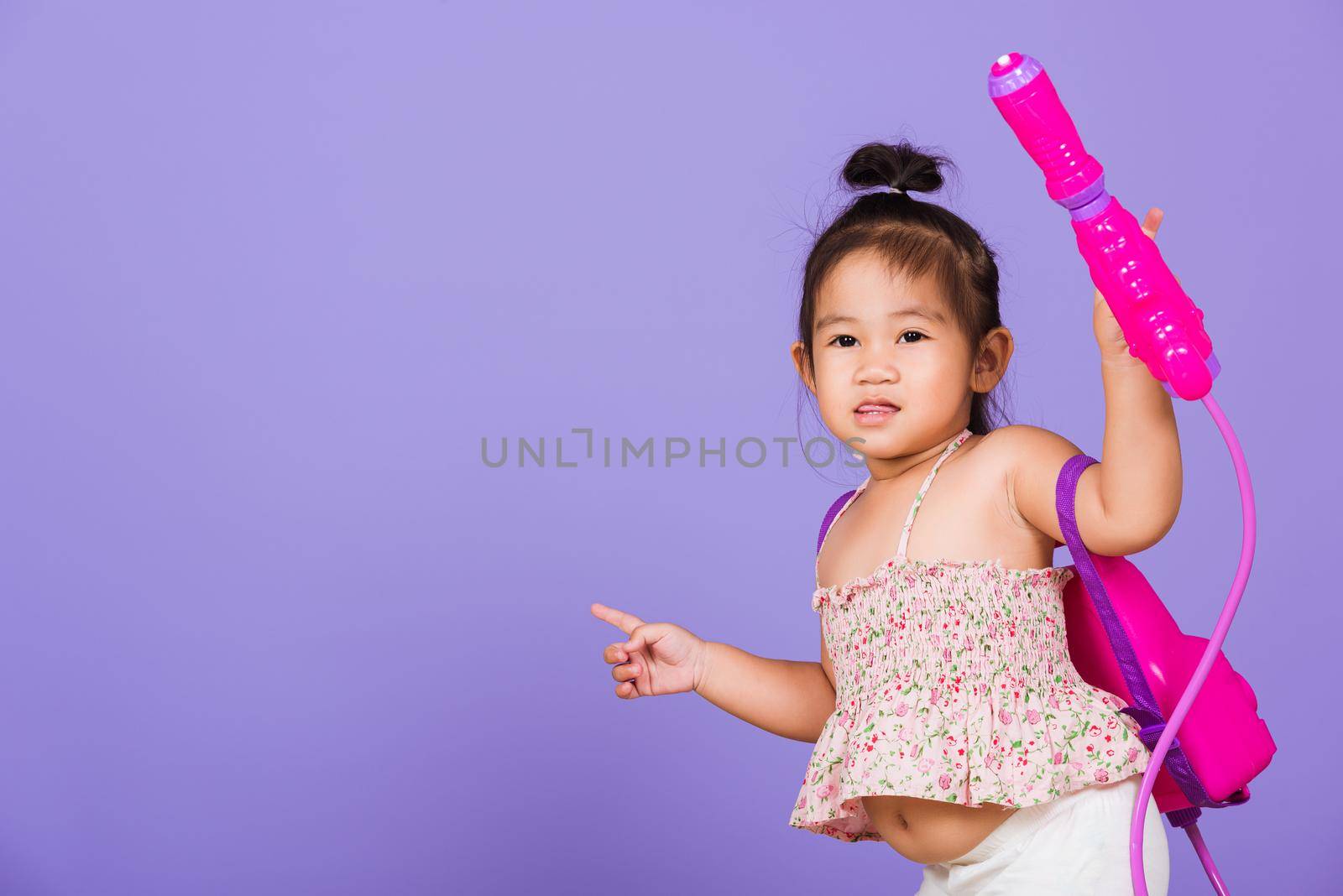 Thai child funny hold toy water pistol and smile, Happy Asian little girl holding plastic water gun, studio shot isolated on purple background, Thailand Songkran festival day national culture concept