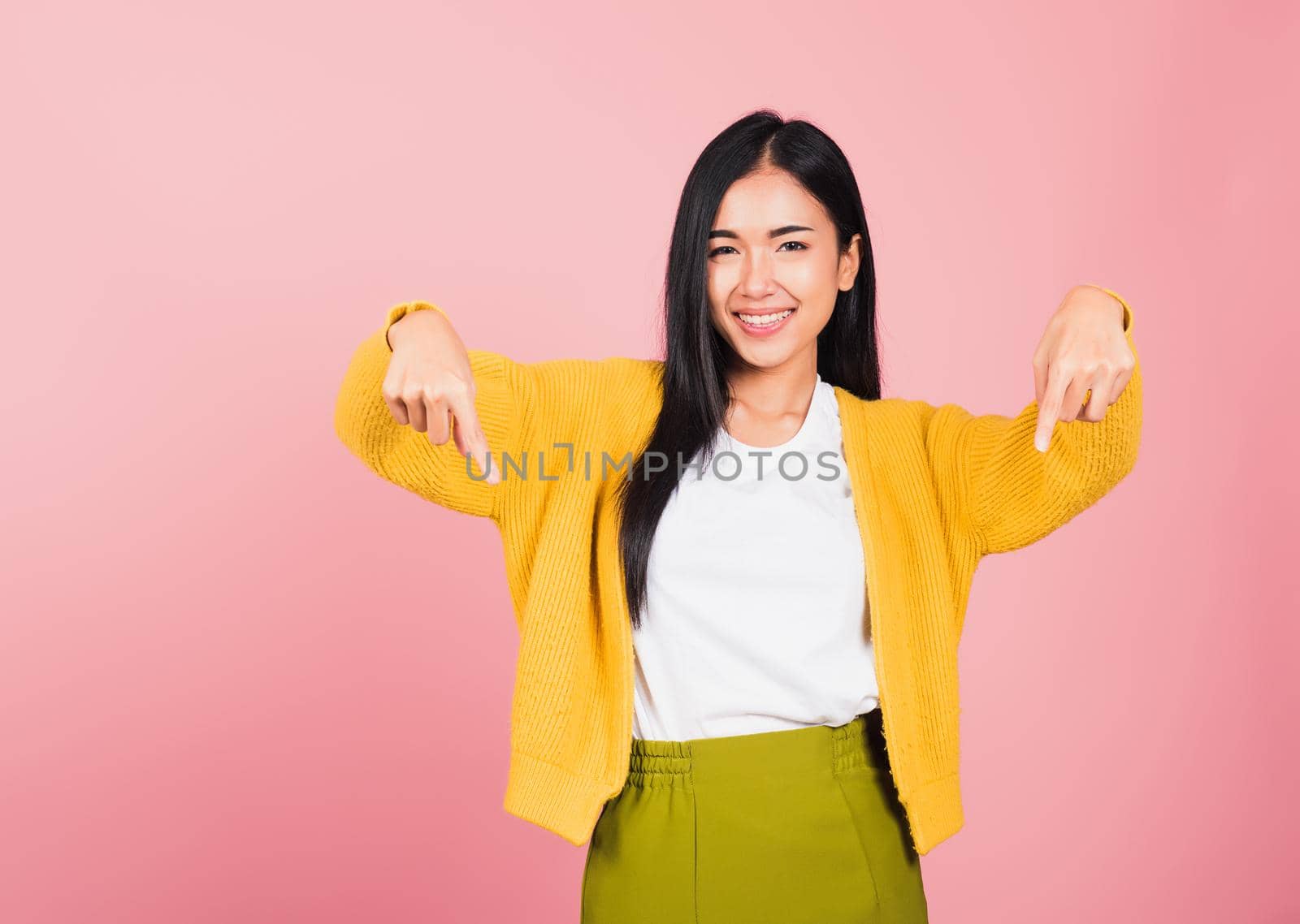 Asian happy portrait beautiful cute young woman smiling stand makes gesture two fingers pointing below down presenting and looking to camera, studio shot isolated on pink background with copy space