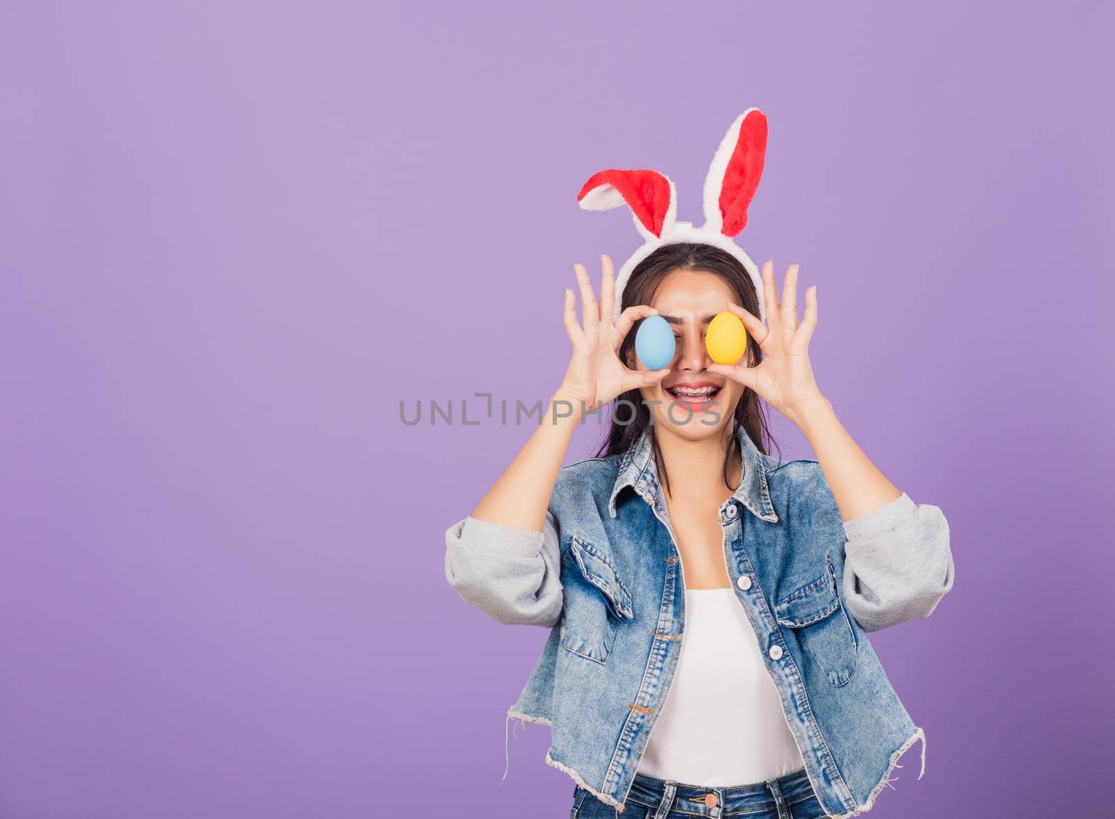 woman smiling wearing rabbit ears and denims holding colorful Easter eggs front eyes by Sorapop