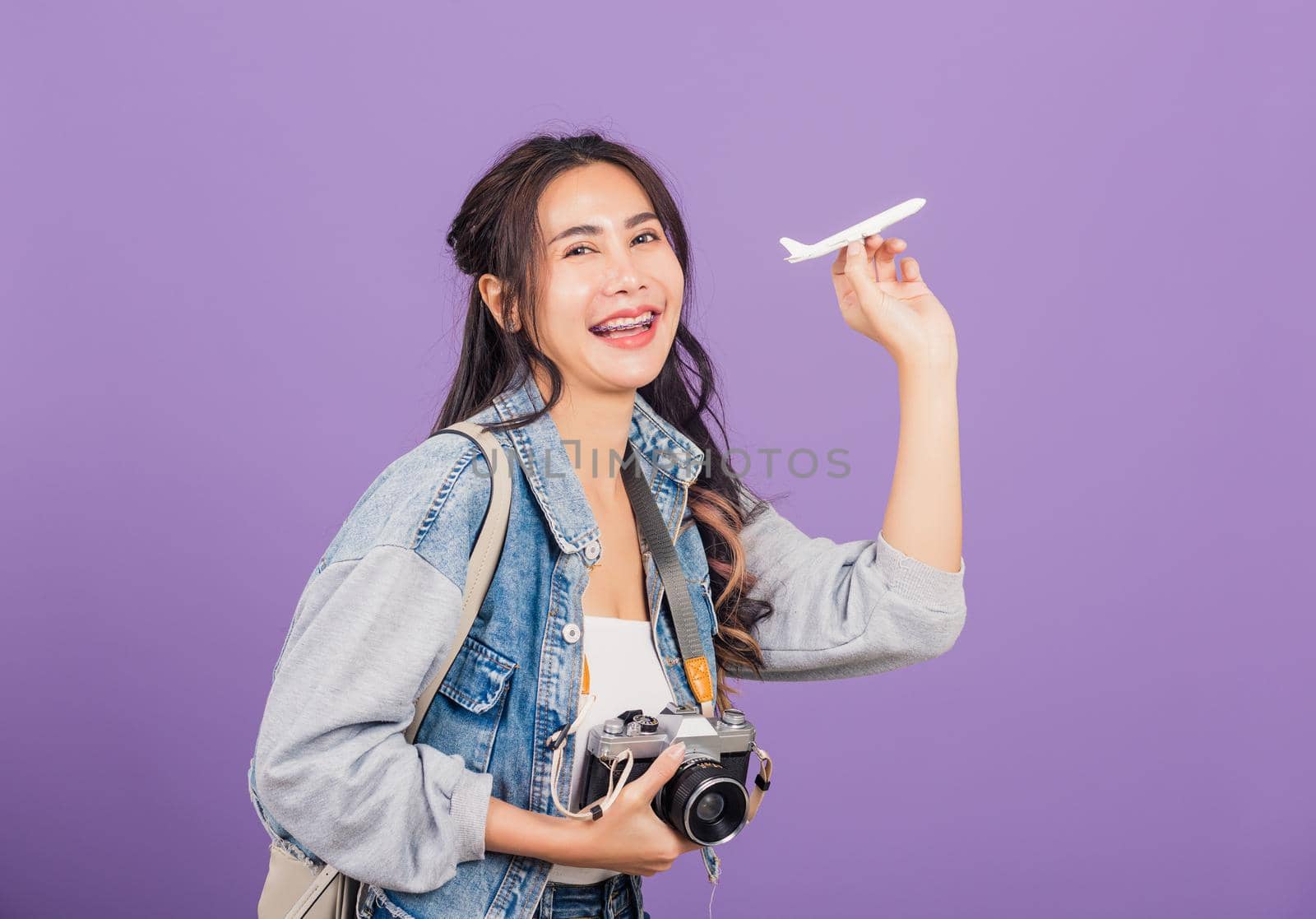 Happy Asian portrait beautiful young woman excited smiling holding airplane toy and retro vintage photo camera, Thai female ready travel trip isolated on purple background, tourism and vacation