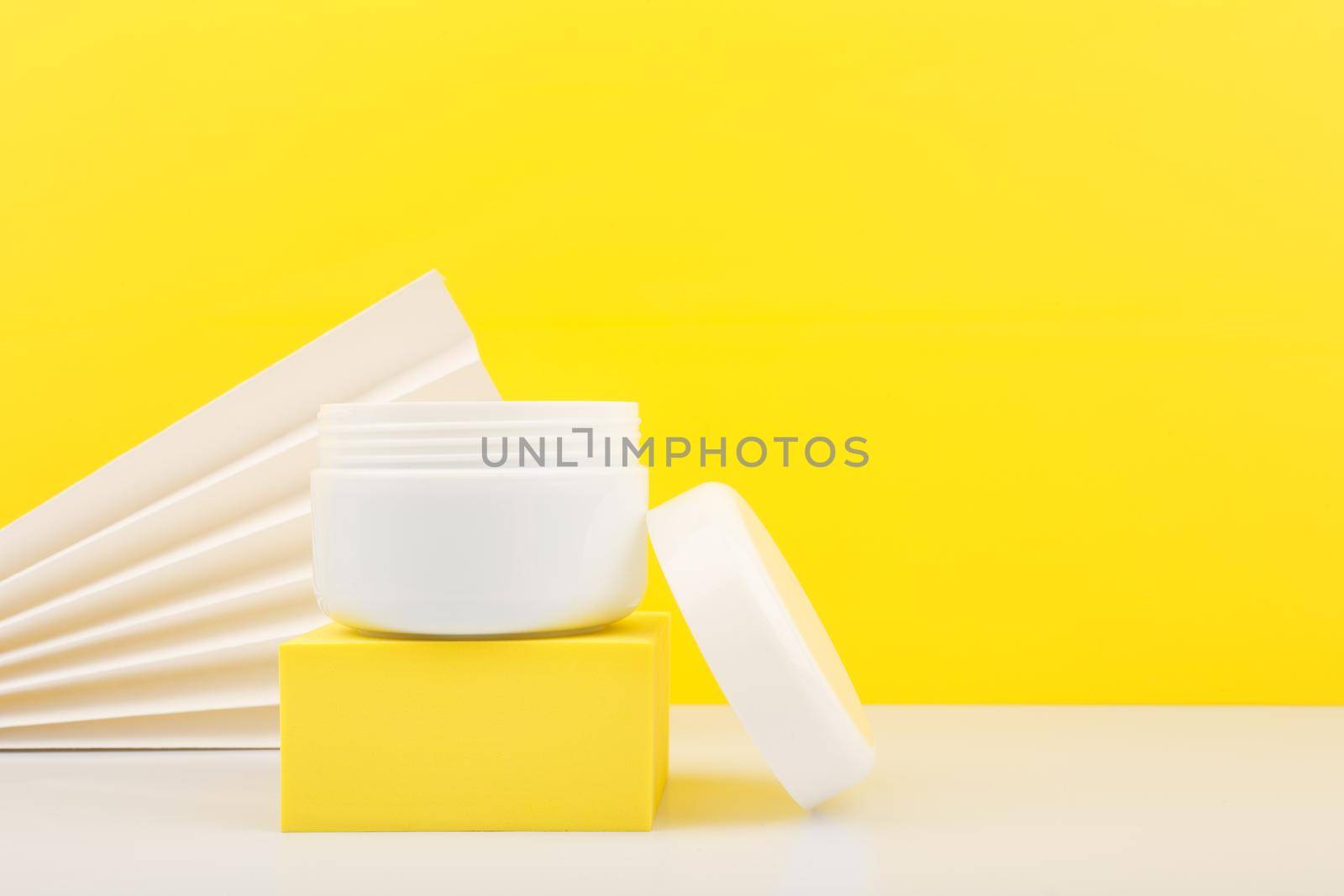 White opened cosmetic jar on yellow podium against yellow background decorated with white waver. Concept of beauty products and skin care