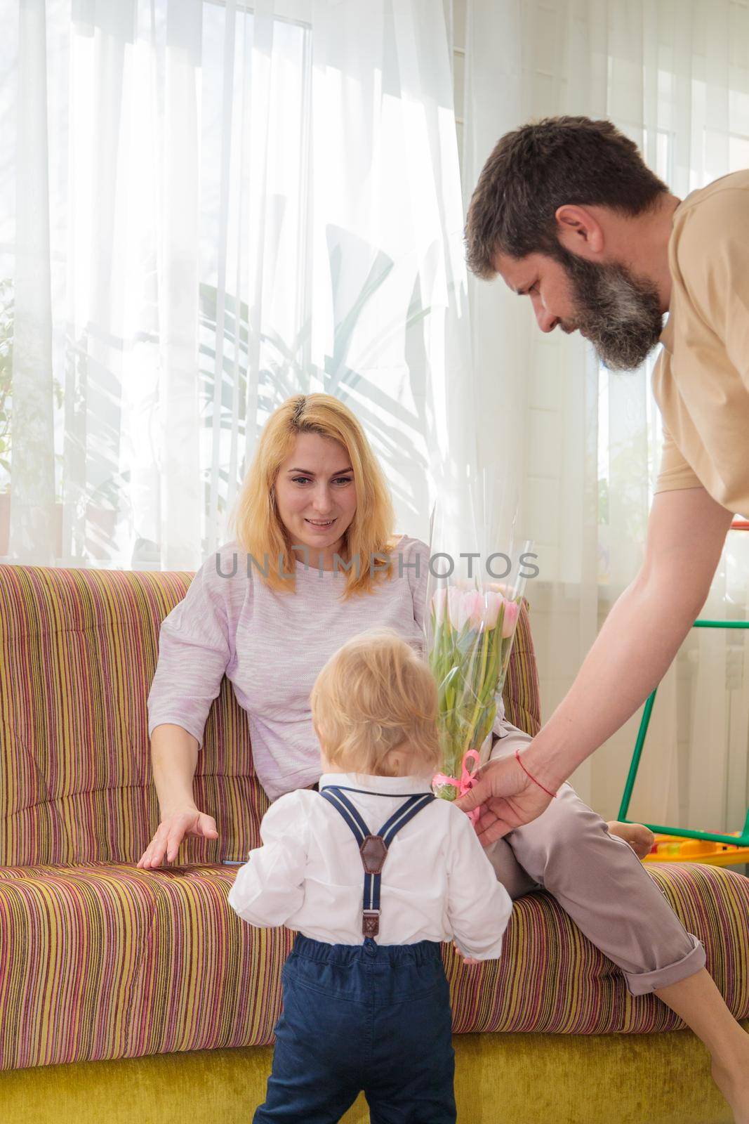 Dad with baby, son congratulate mom on the holiday. The kid brings his mother a bouquet of flowers and a gift.