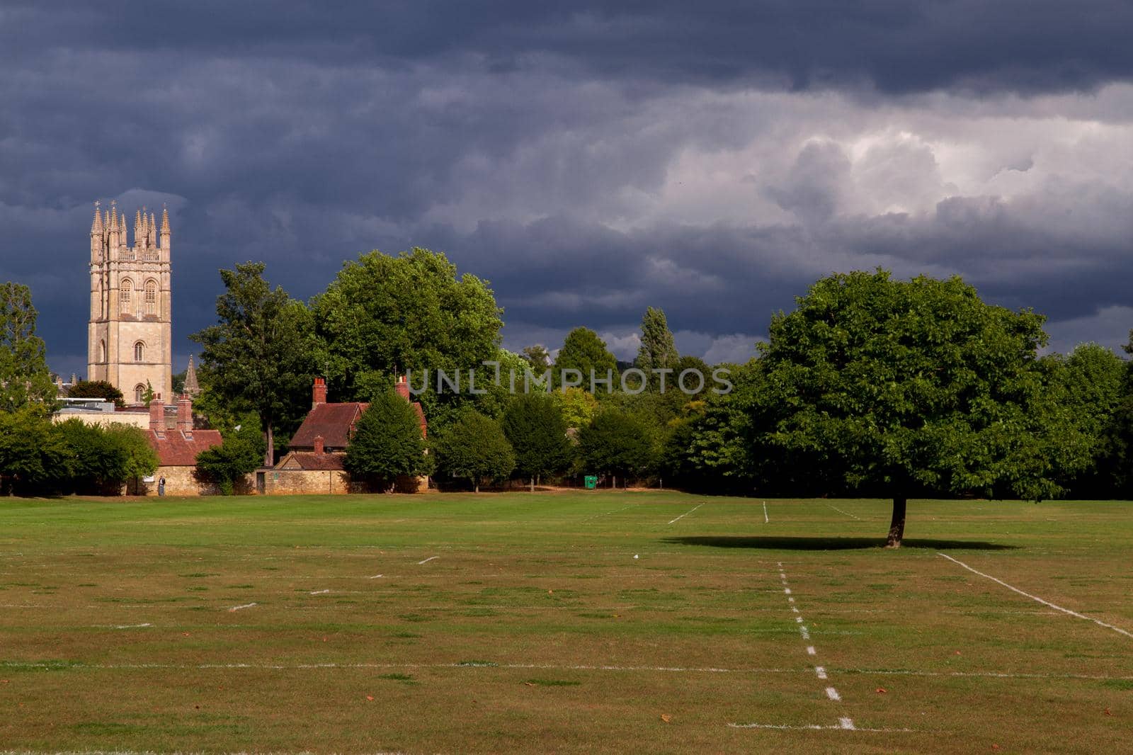 Wide-shot of a sport field surrounded by trees and some small houses and church tower in the background on a sunny day with a cloudy sky