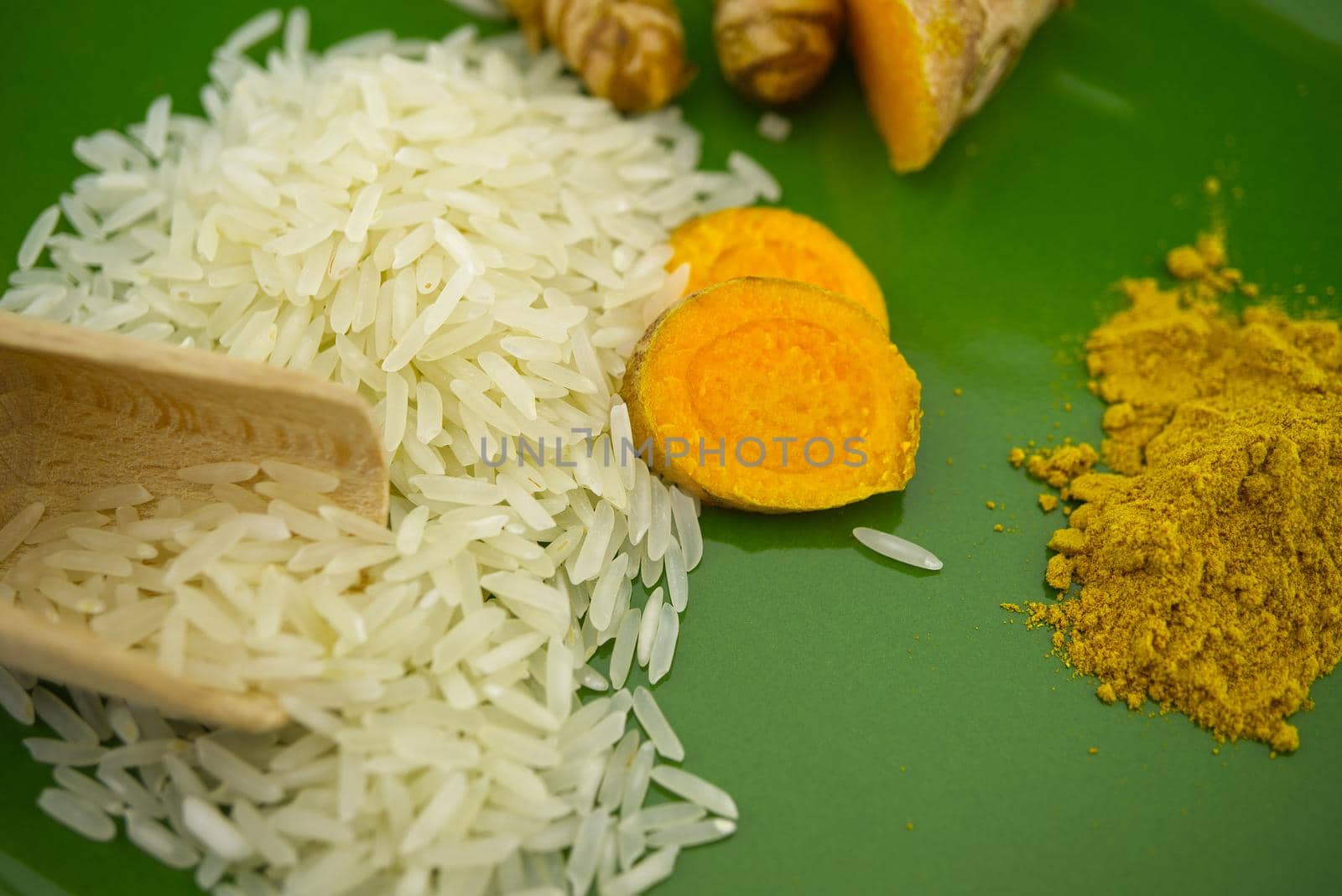 Turmeric roots and slices next to heaps of rice and spice powder on a green plate. Selective focus and free space for text