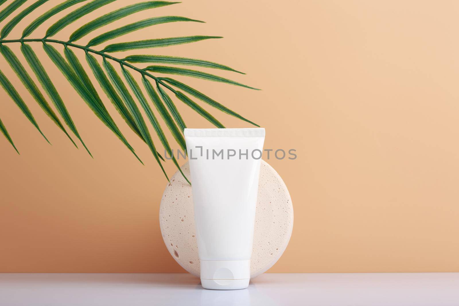 White cosmetic tube against round gypsum podium with palm leaf against beige background with copy space. Concept of beauty and skin care. Unbranded tube with cream, lotion, gel or foam for skin care