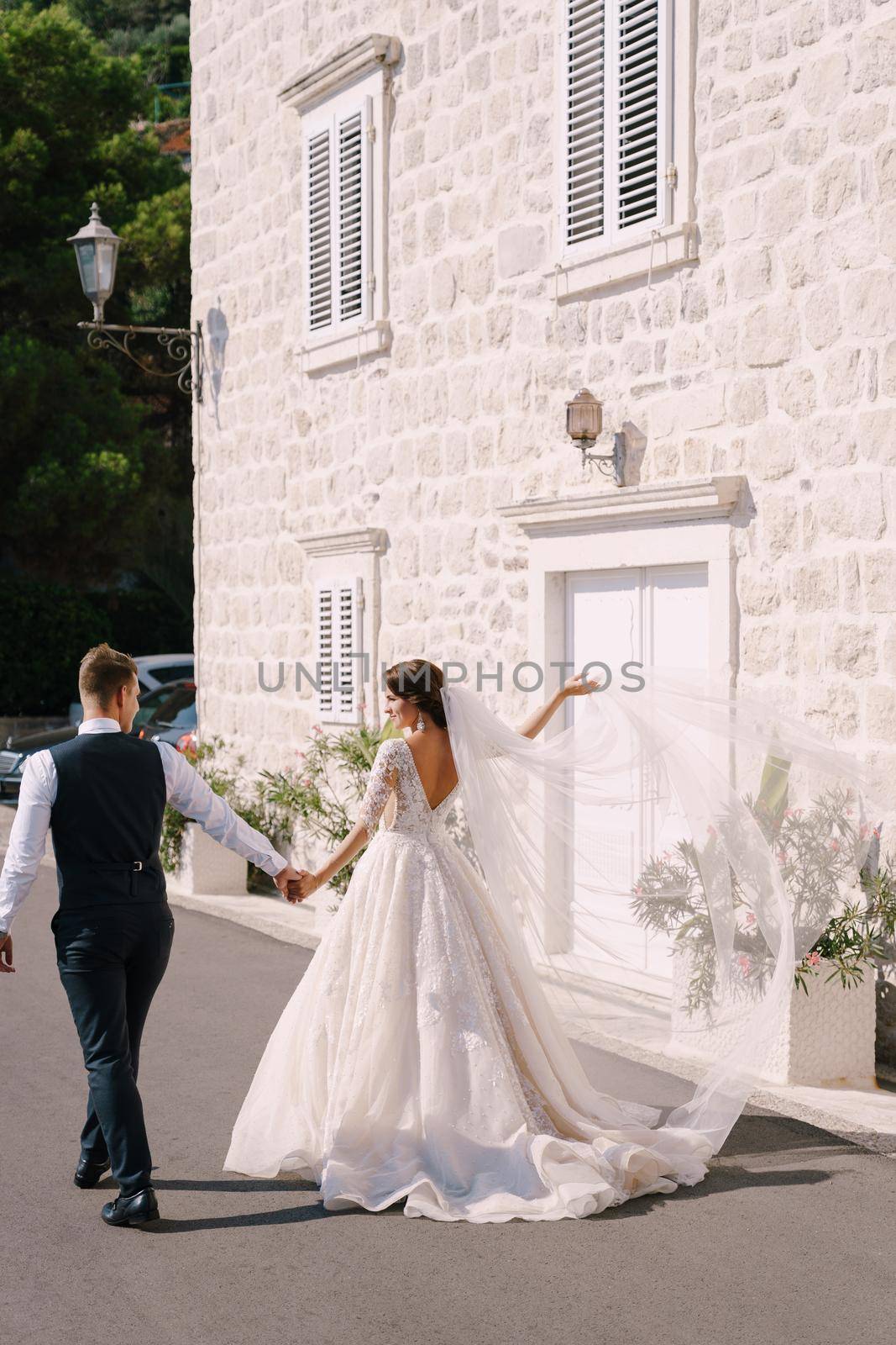 A wedding couple walks along the promenade against the background of a white old house, the bride waves a long veil, the groom holds her hand. Fine-art wedding photo in Montenegro, Perast. by Nadtochiy
