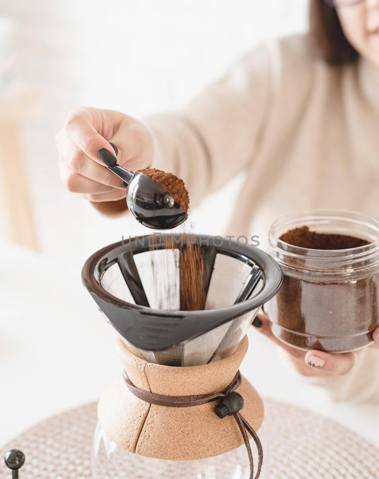 Alternative coffee brewing. Young woman brewing coffee in chemex sitting at the white table with various stuff for alternative coffee brewing, pouring grinded coffee beans into filter