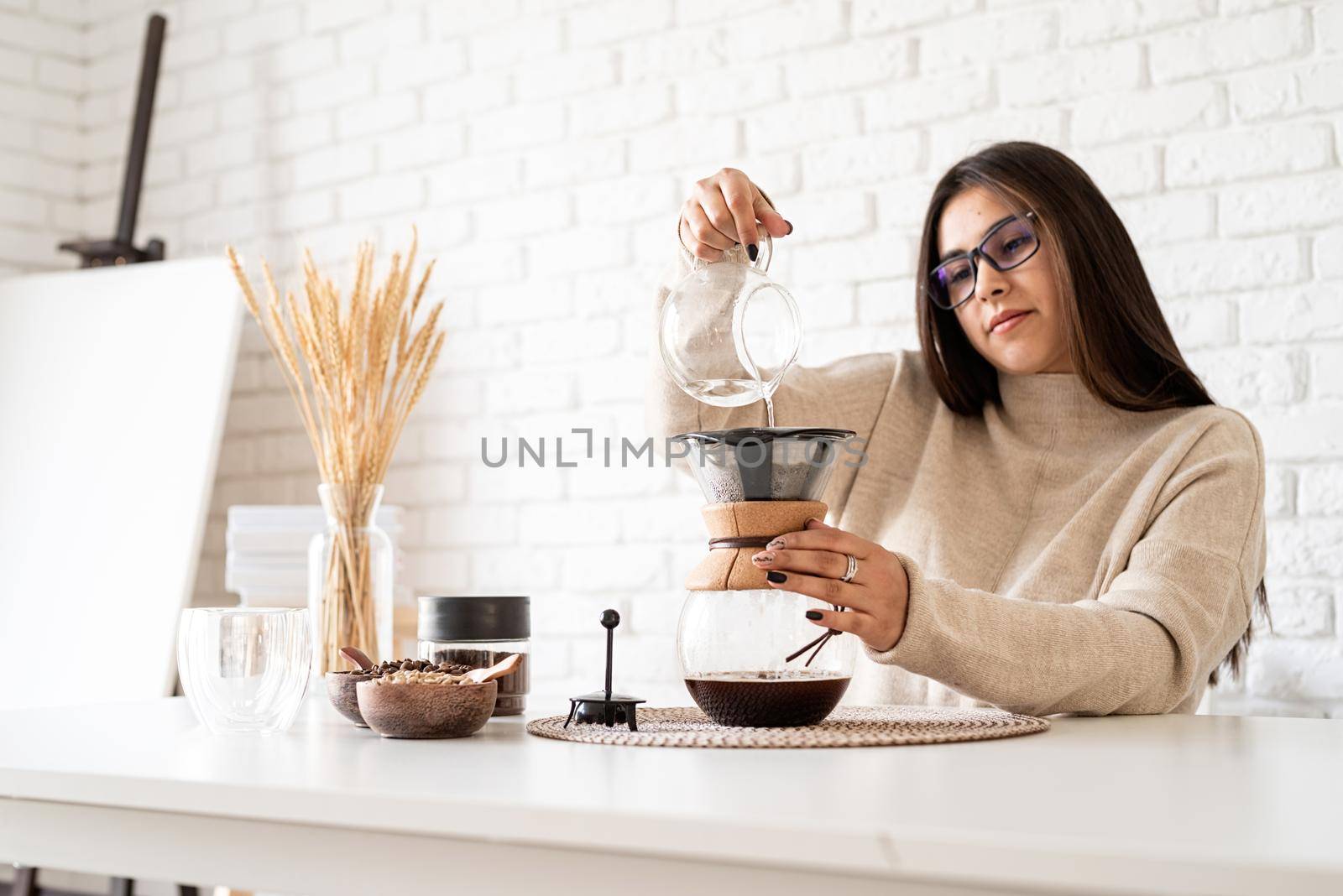 Alternative coffee brewing. Young woman brewing coffee in chemex standing at the white table with various stuff for alternative coffee brewing, pouring hot water into the filter