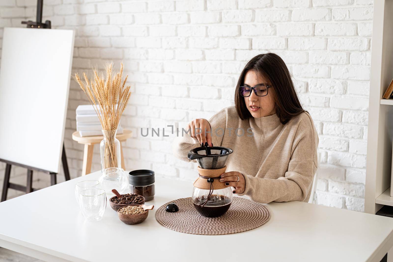 Alternative coffee brewing. Young woman brewing coffee in chemex standing at the white table with various stuff for alternative coffee brewing, pouring hot water into the filter
